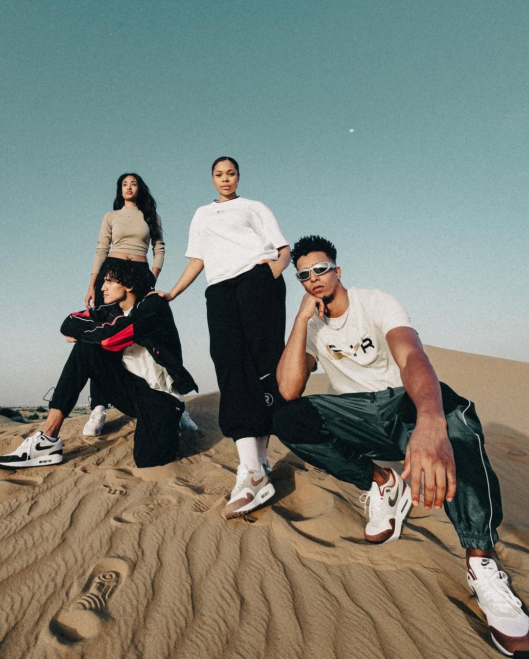 Four young individuals in casual and athletic clothing posing on sand dunes under a clear sky, with one person sitting and two standing, and another crouching.