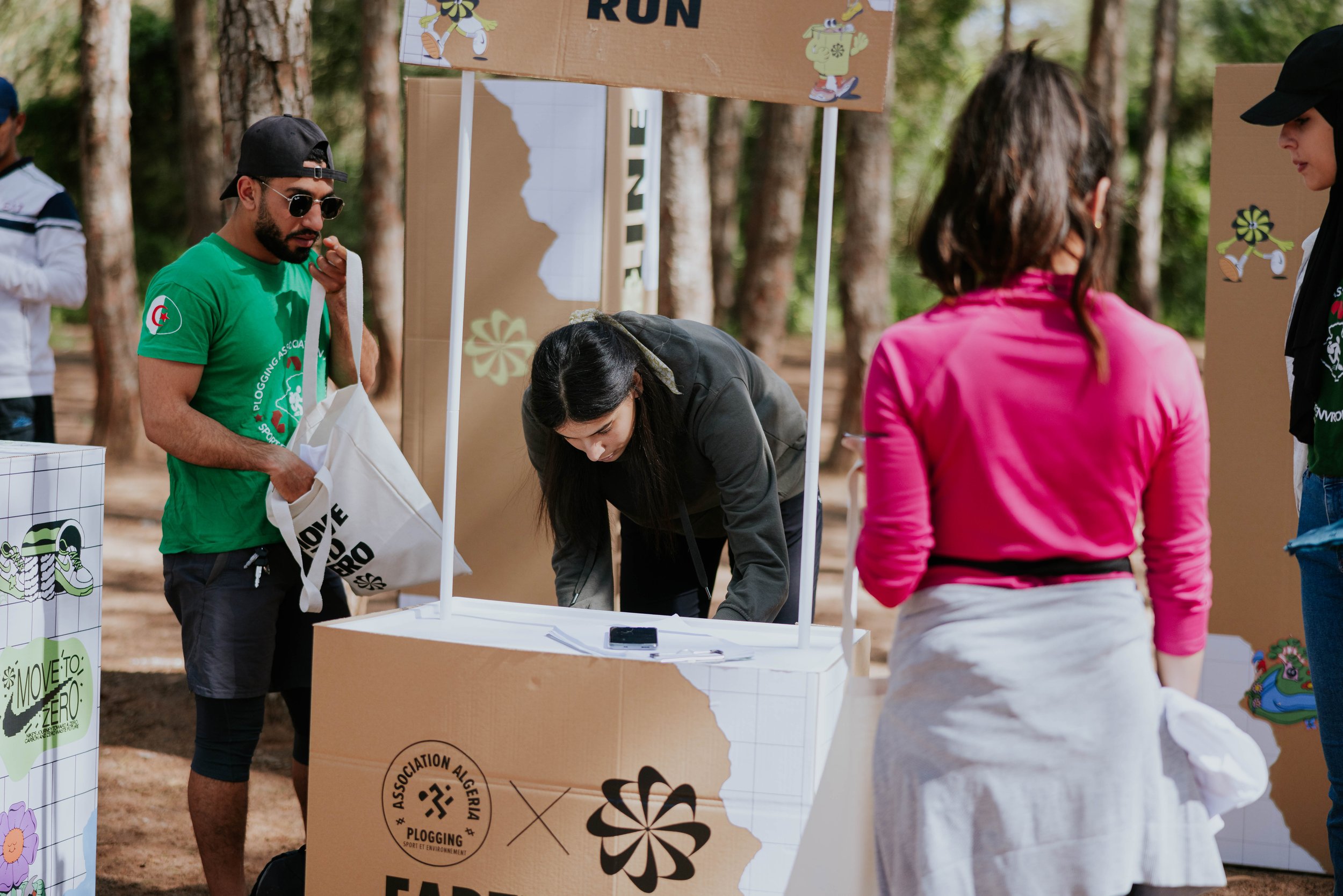 People at an outdoor event with booths and banners, some are engaging with displays, in a wooded area with trees in the background.