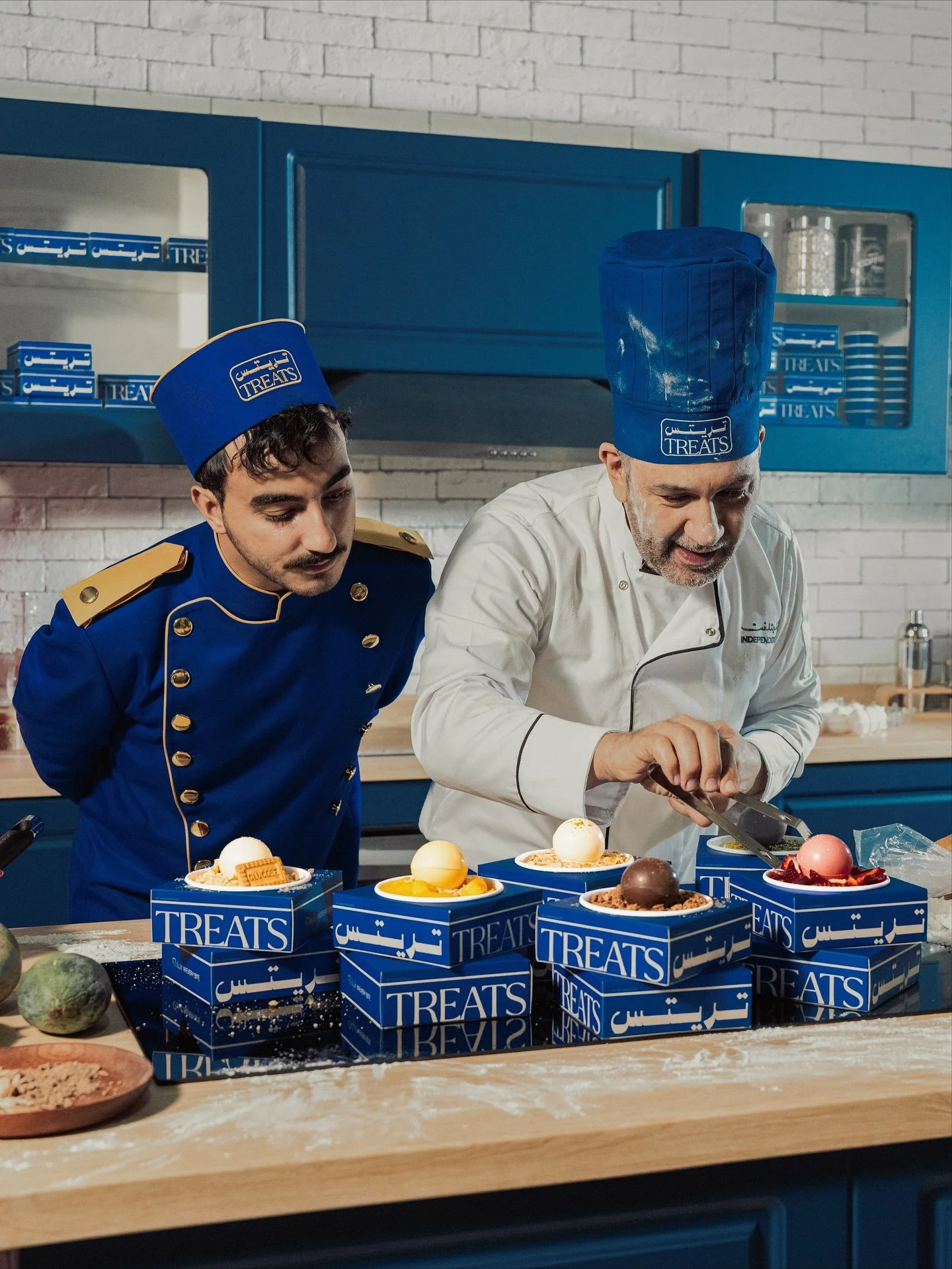 Two chefs in blue and white uniforms decorating cakes in a modern kitchen with white brick walls and blue cabinets. Several blue boxes labeled "TREATS" with cakes and desserts inside are on the table.