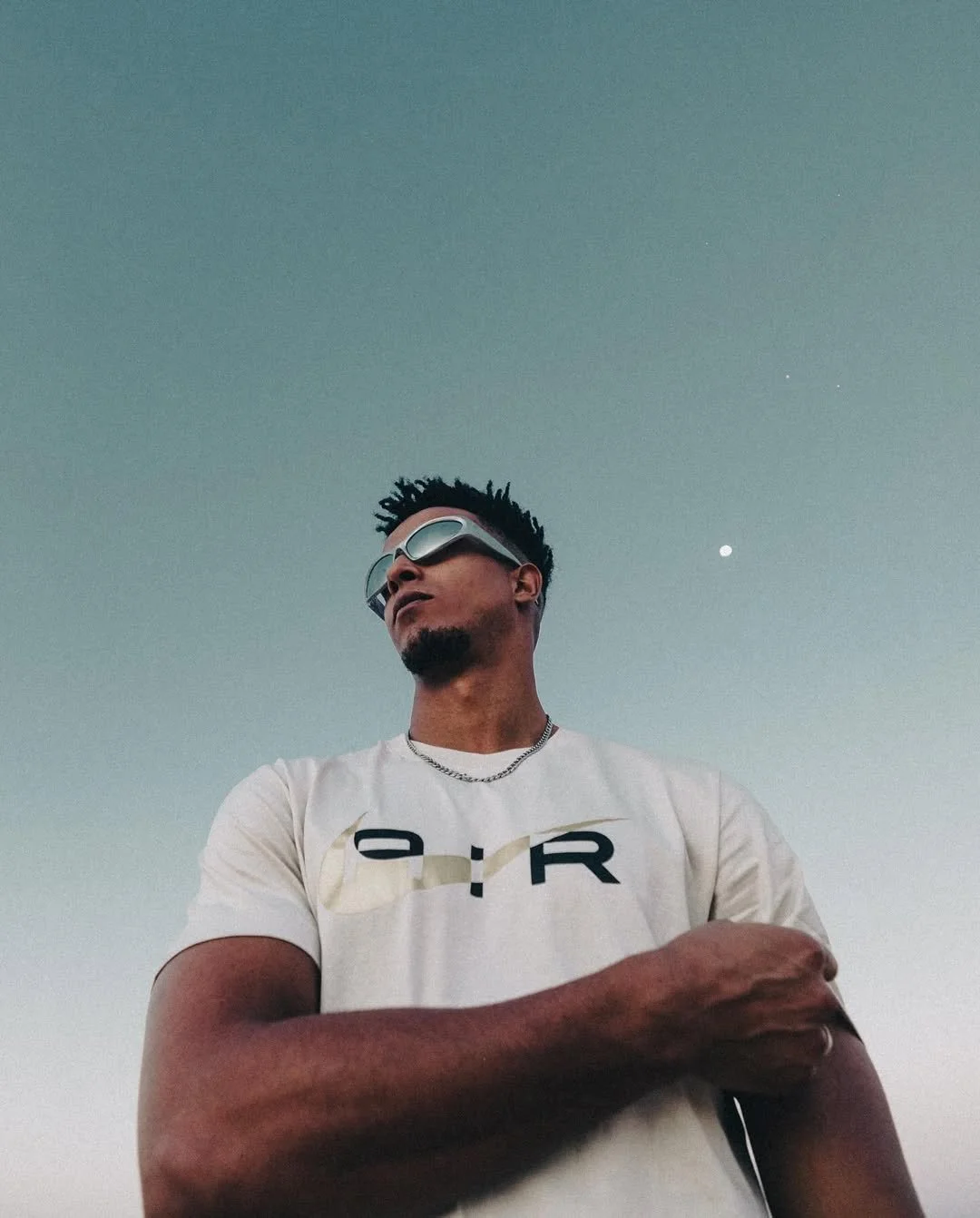 A man wearing sunglasses and a white graphic t-shirt standing outdoors against a clear sky with the moon visible in the background.