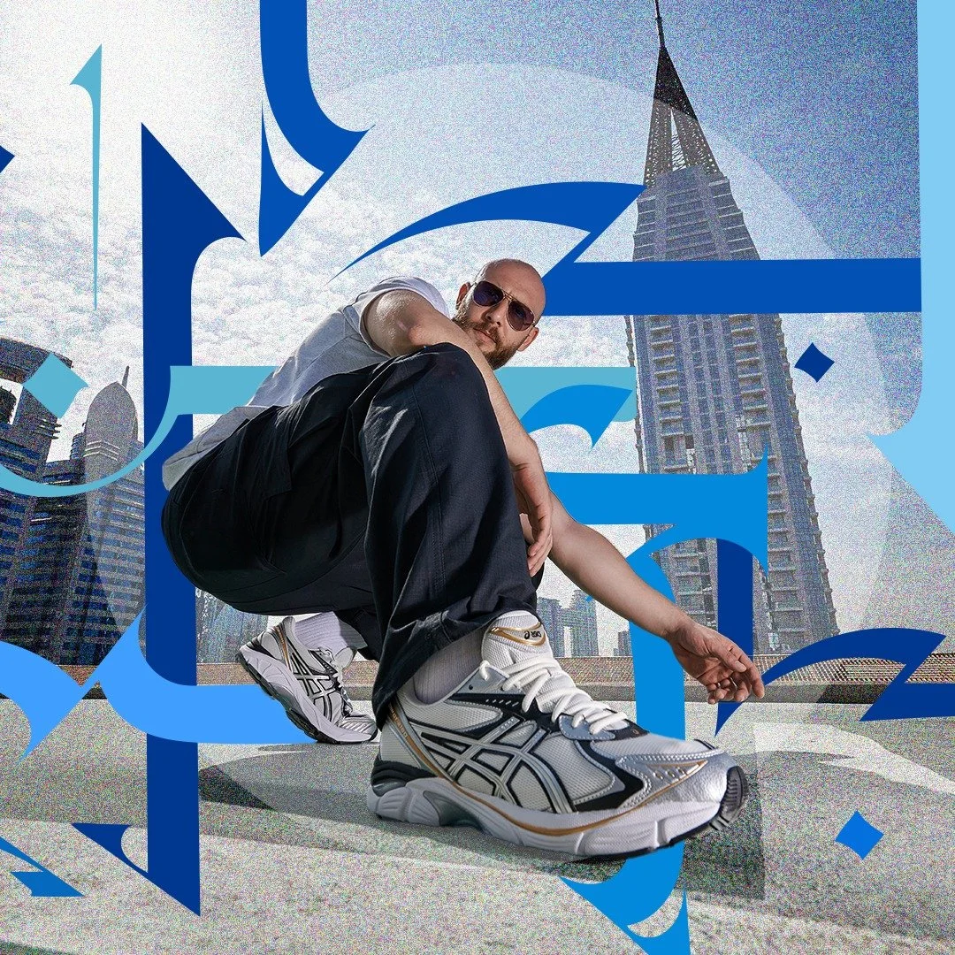 A man with a bald head, sunglasses, a white shirt, black pants, and white sneakers crouches on a rooftop with a city skyline, including the Empire State Building, in the background. The image has blue graphic elements overlaid.
