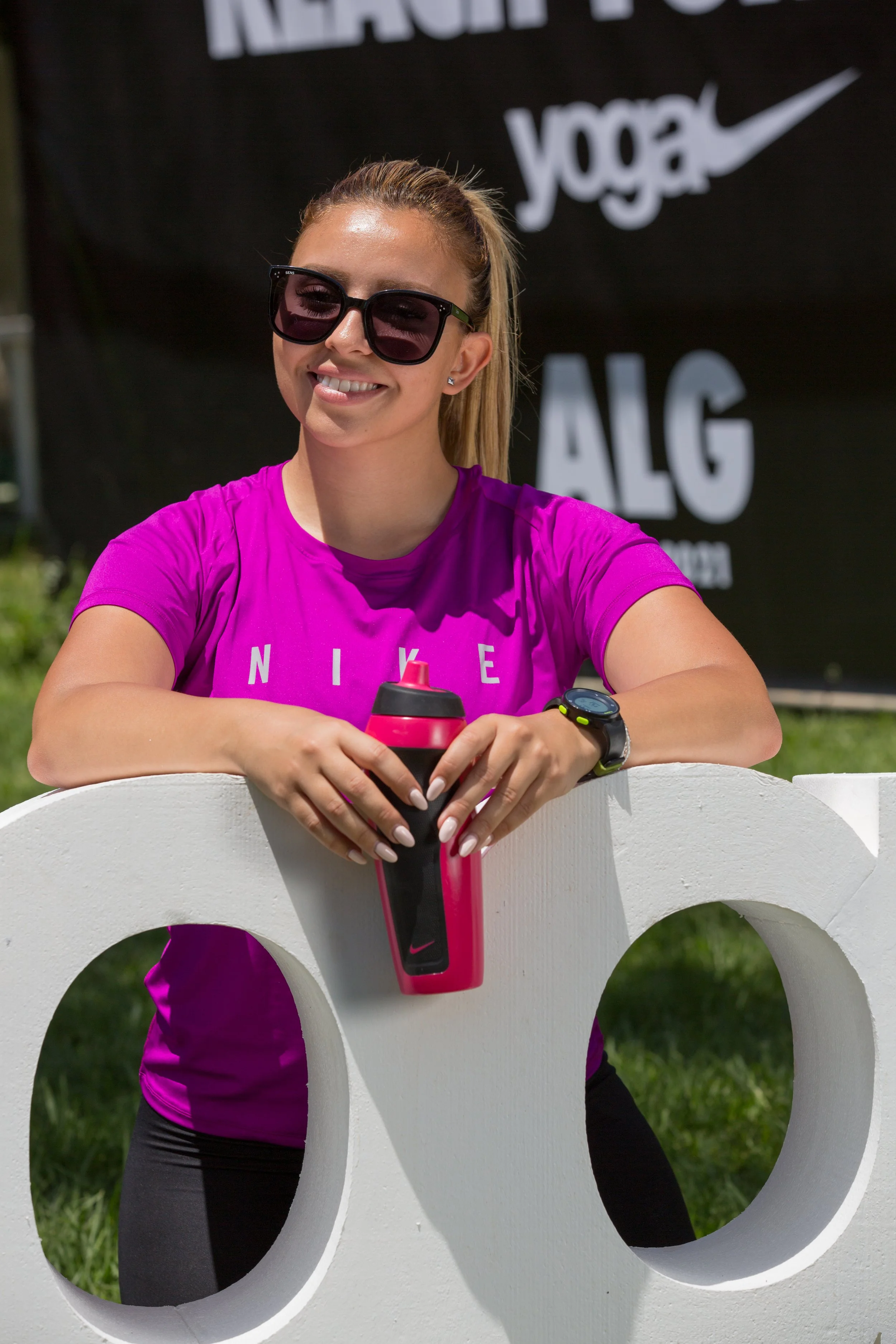 A smiling woman wearing black sunglasses and a bright pink Nike T-shirt, holding a pink and black water bottle, standing behind a decorative white fence with circular cutouts, outdoors with green grass and a large black Nike sign in the background.