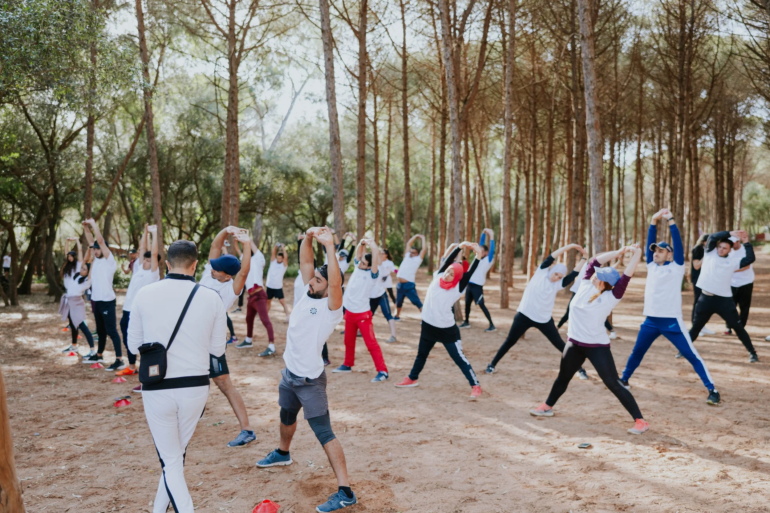 A large group of people participating in an outdoor exercise session in a forested area. The participants are stretching or doing yoga poses, wearing athletic clothing, and are spread out across a dirt clearing among tall trees.