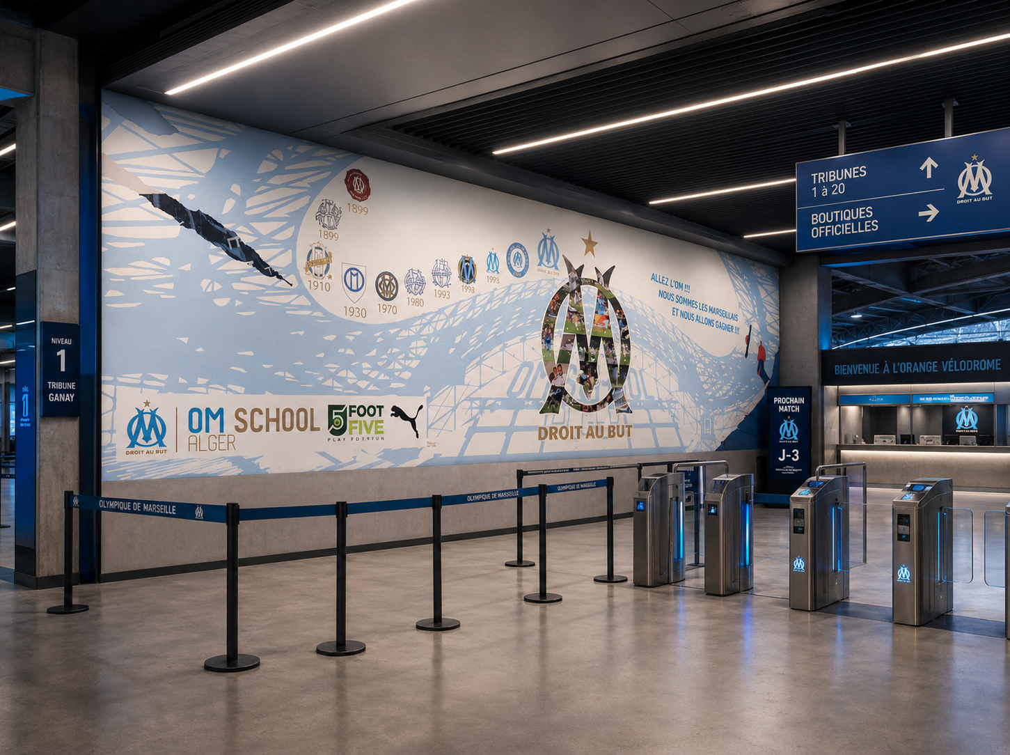 Interior of Marseille football stadium with large mural celebrating the club's history, logos, and achievements, ticket barriers at front, signage indicating entrance directions, and a reception area behind.