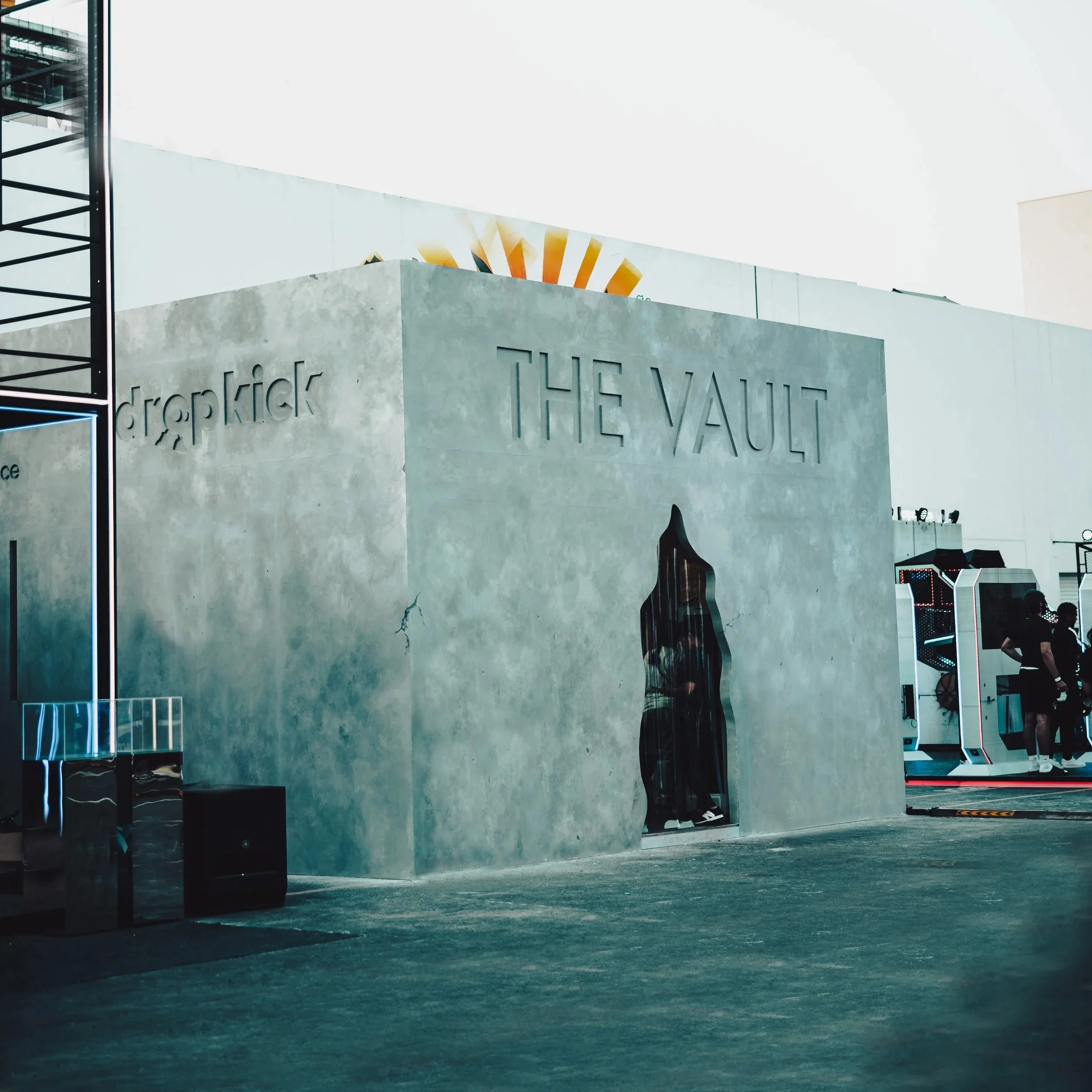 A modern, concrete building with the words 'THE VAULT' and 'dropkick' engraved on it, with colorful signs and advertisements nearby, and an overcast sky in the background.