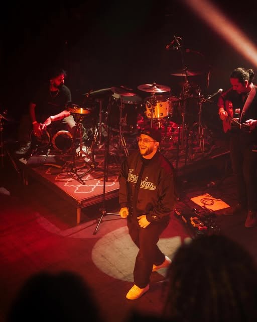 A musician in a black hoodie with 'Louisiana' written on it, smiling and standing on stage with a band setup including drums and other instruments in the background.