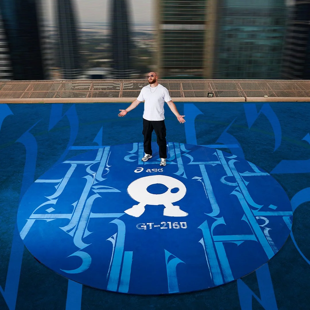 A man with sunglasses and a white t-shirt stands with arms outstretched on a rooftop basketball court with a blue graphic design and a white logo in the center. Tall buildings are visible in the background.