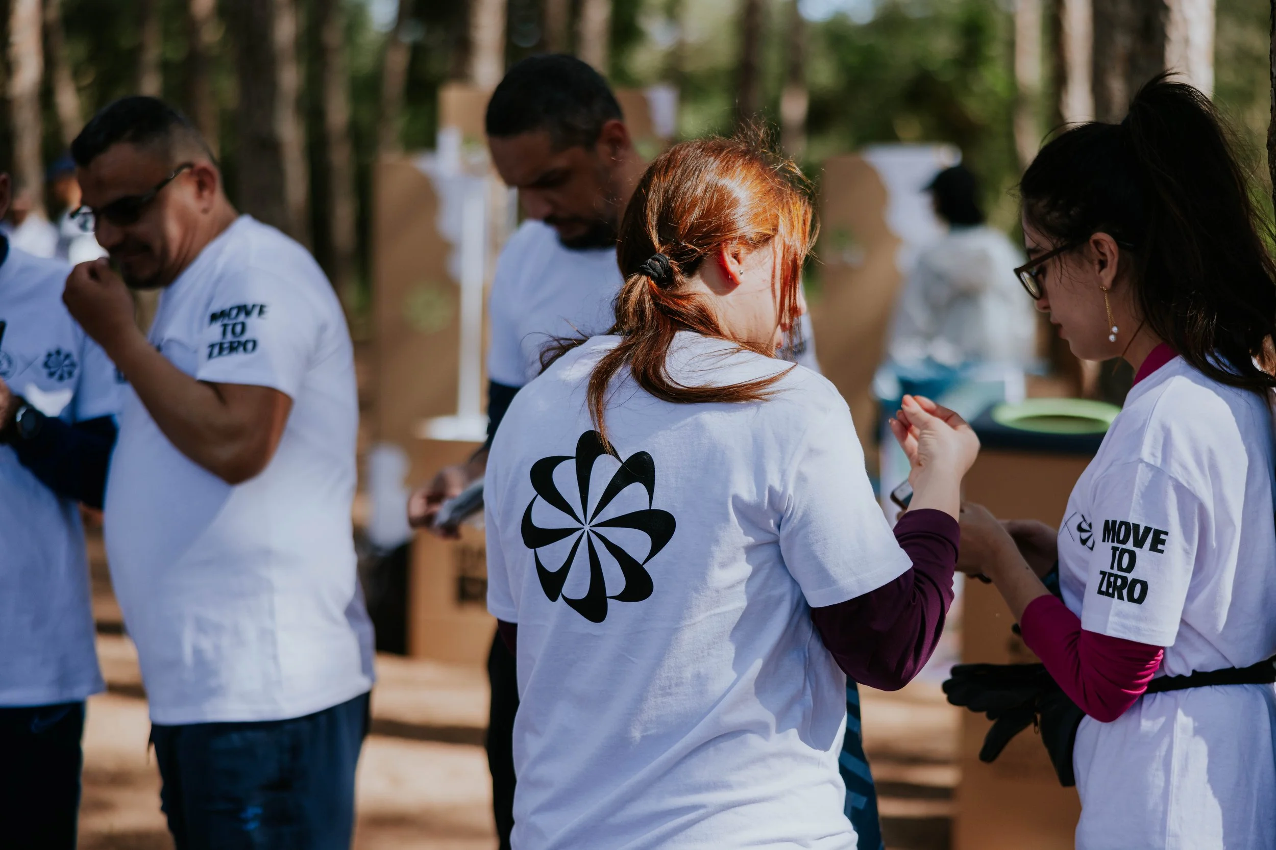 Group of people standing outdoors in a circle, wearing white shirts with a black flower symbol and the phrase "Move to Zero," possibly participating in an event or activity in a wooded area.