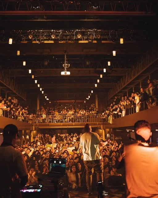 A performer on stage in front of a large crowd in a concert venue, with people on the upper level watching from balconies.