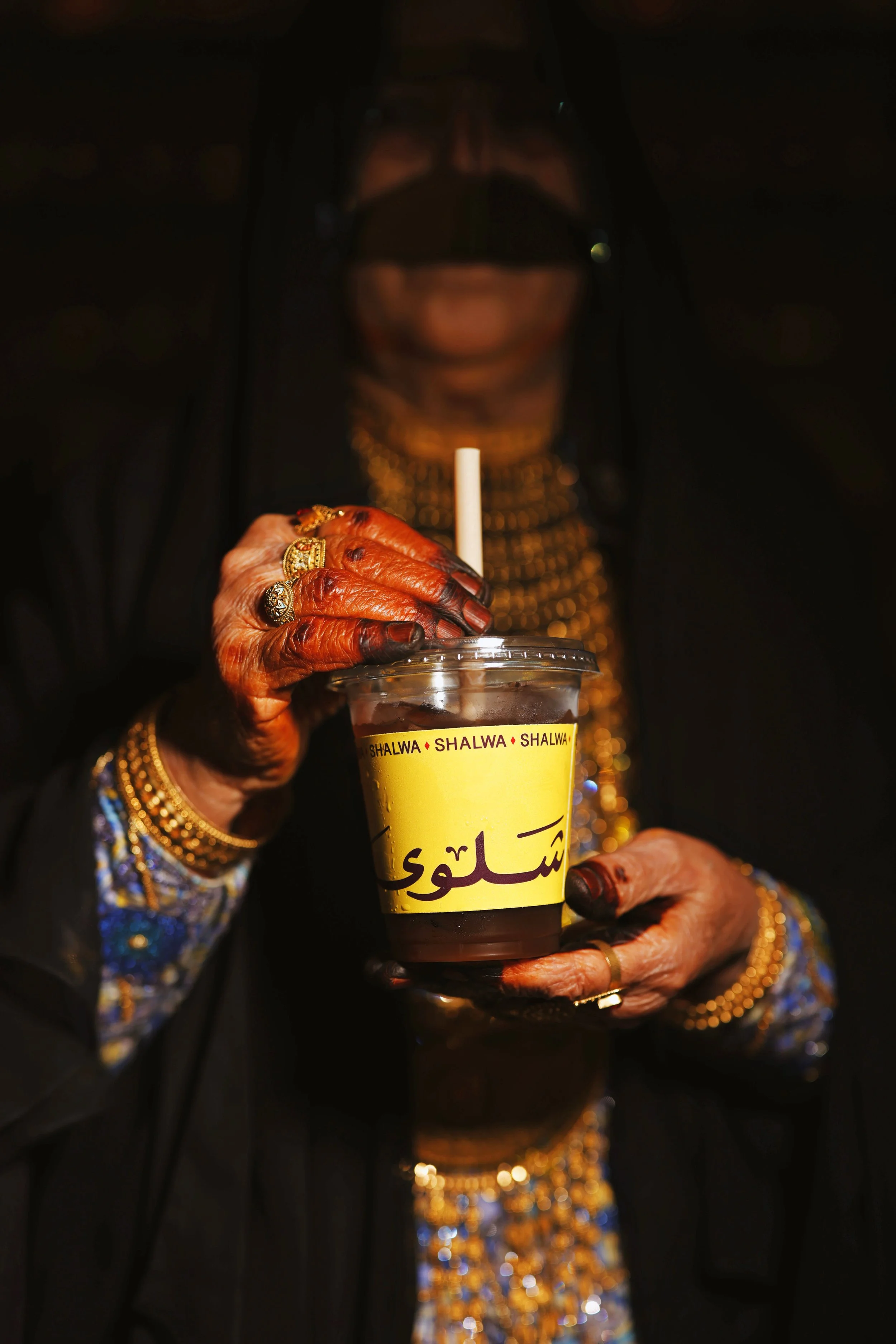 An elderly person with dark skin and jewelry, wearing a black head covering, is holding a yellow cold drink cup with Arabic writing on it. The person's face is blurred, and their hand and fingers are adorned with multiple rings and gold bangles. The person is sipping the drink through a straw.