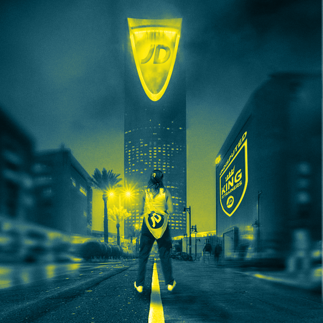 A person standing on a city street at night, with illuminated signs for 'JD' and 'KING TRAINERS', and tall buildings with lit windows in the background.
