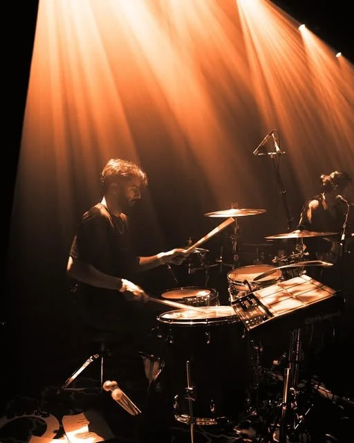 Musicians playing drums on stage with orange lighting.