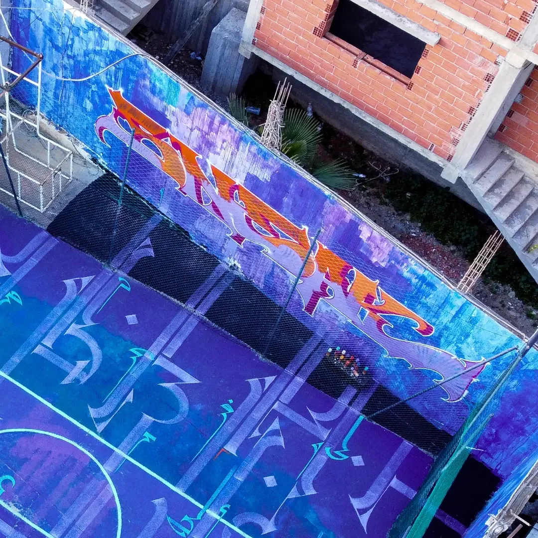 An outdoor basketball court with a colorful, artistic floor design and a large graffiti-style mural on a wall. The court is enclosed by a high fence, and there are construction buildings in the background.
