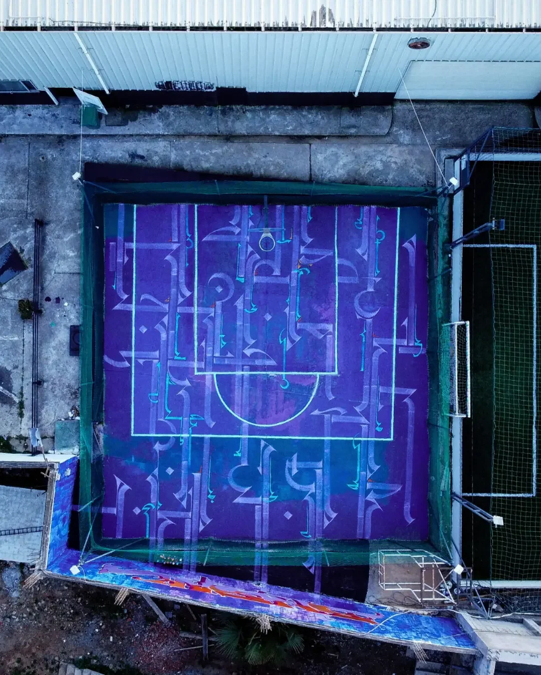 An aerial view of a basketball court with artistic purple markings under UV light, surrounded by a green fence.
