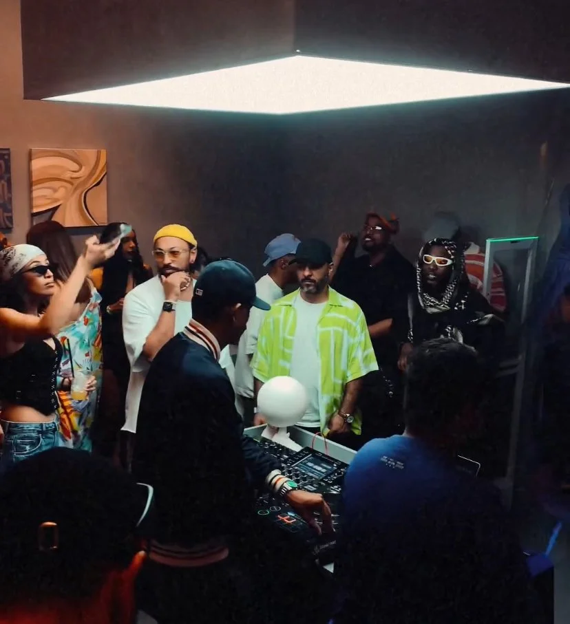 Group of young people dancing and socializing in a dimly lit room with a DJ playing music.