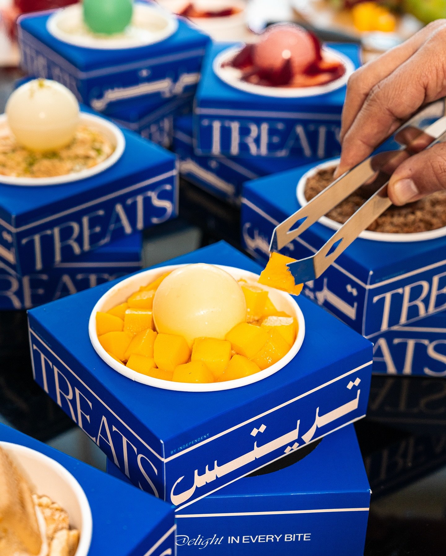 Close-up of Blue boxes containing frozen desserts topped with fruits like mango and peach, with someone using tongs to select a portion.