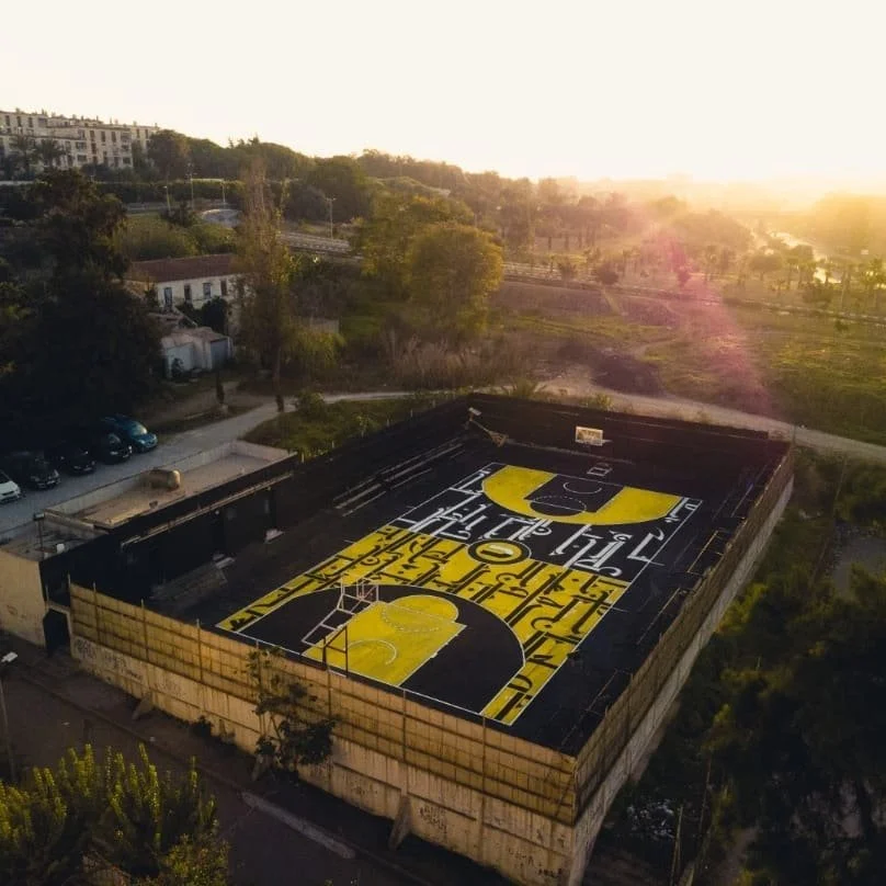 A basketball court on top of a building with a yellow and black design, seen from above during sunset with trees, roads, and buildings surrounding it.
