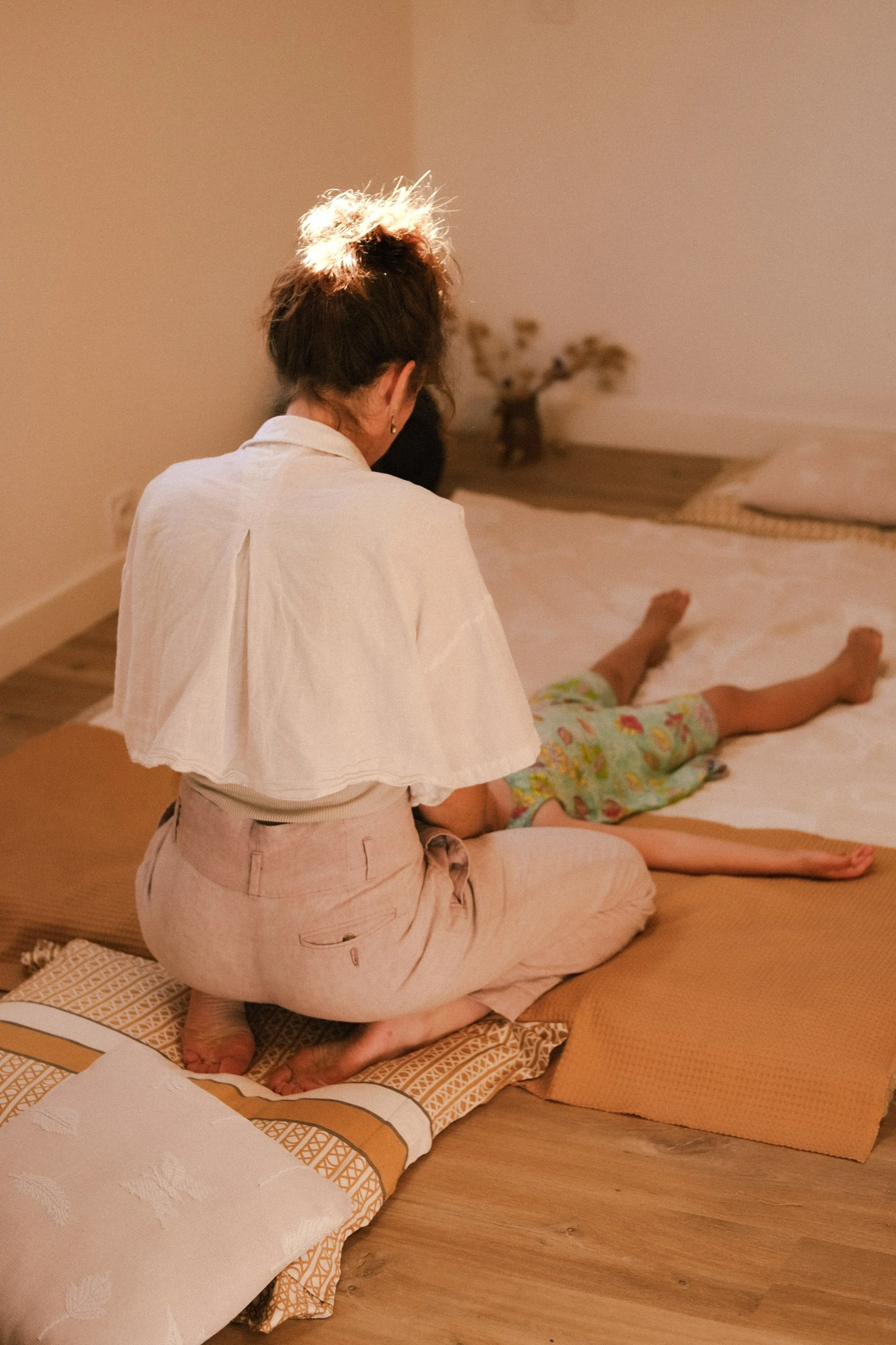 Une femme est assise à genoux sur un tapis, regardant vers une enfant allongée sur un matelas avec un pyjama coloré. La scène se passe dans une pièce chaleureuse au sol en bois, avec des coussins et un lit en arrière-plan.