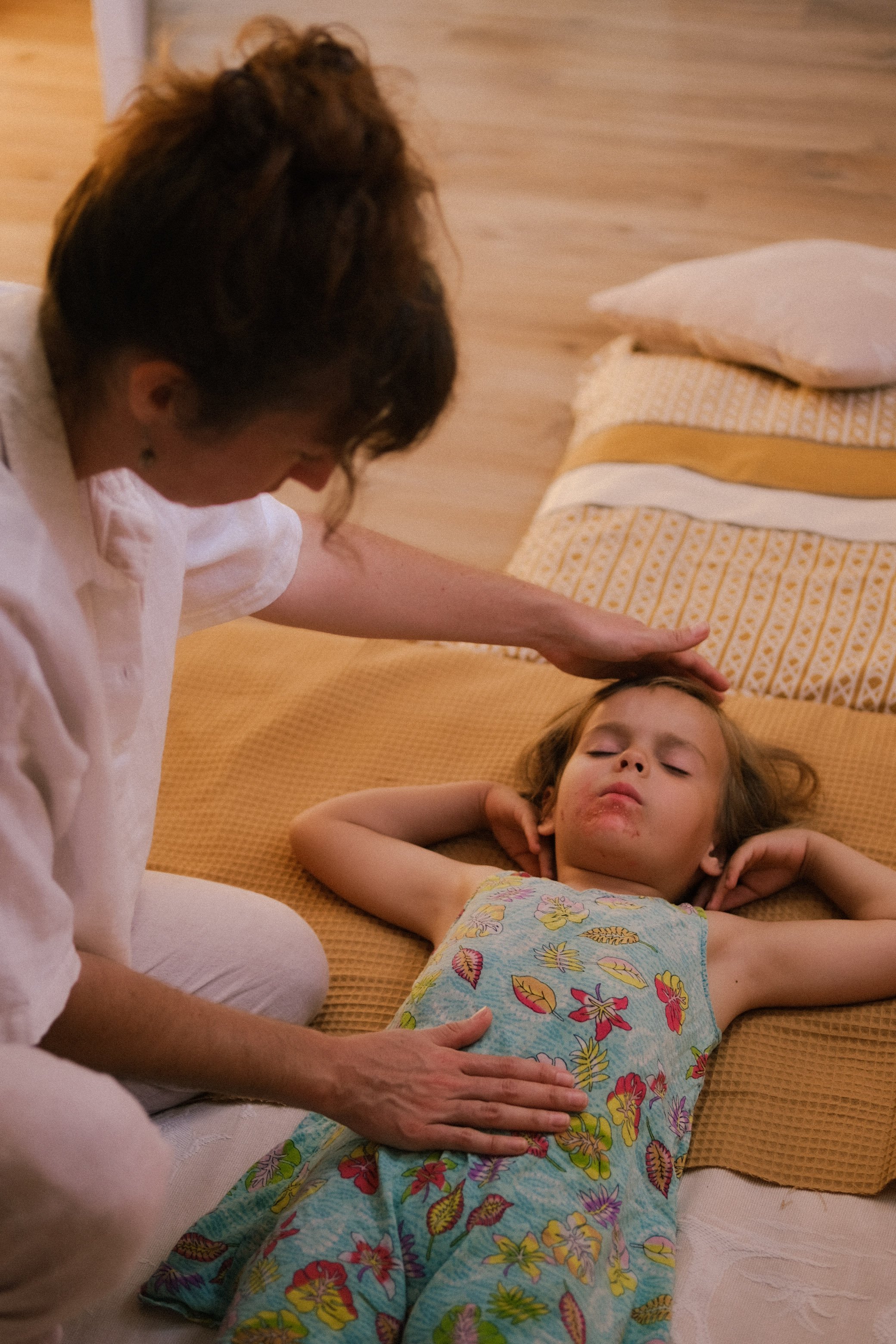Une femme semble faire une séance de rééducation ou de relaxation sur une fillette allongée sur un lit ou un matelas. La femme est agenouillée, avec la main sur le ventre de la fillette, qui est allongée, yeux fermés, vêtue d'une robe colorée avec des fleurs.
