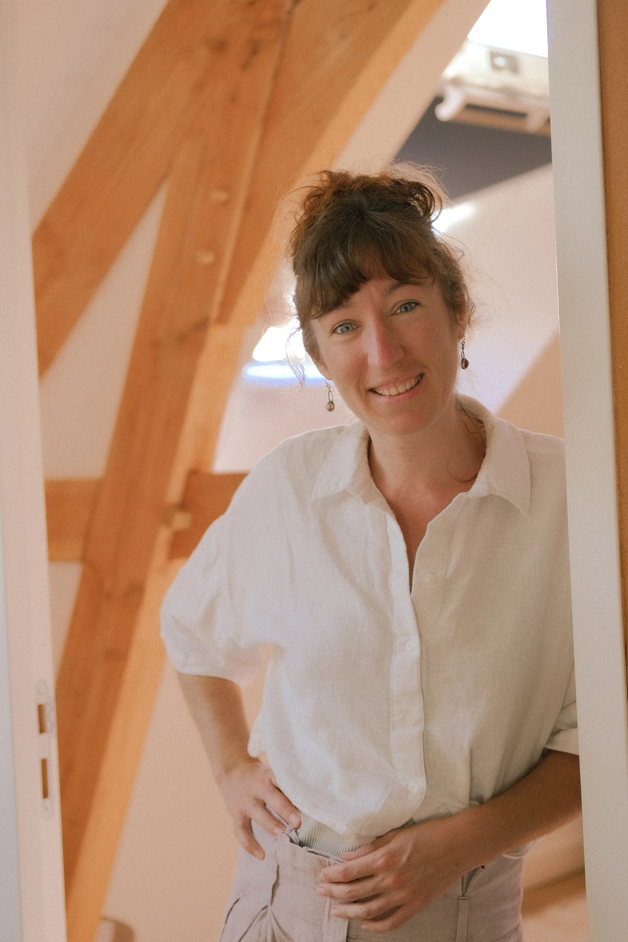 Une femme souriante portant une chemise blanche et un pantalon beige, prise dans le miroir d'une pièce avec des poutres en bois au plafond.