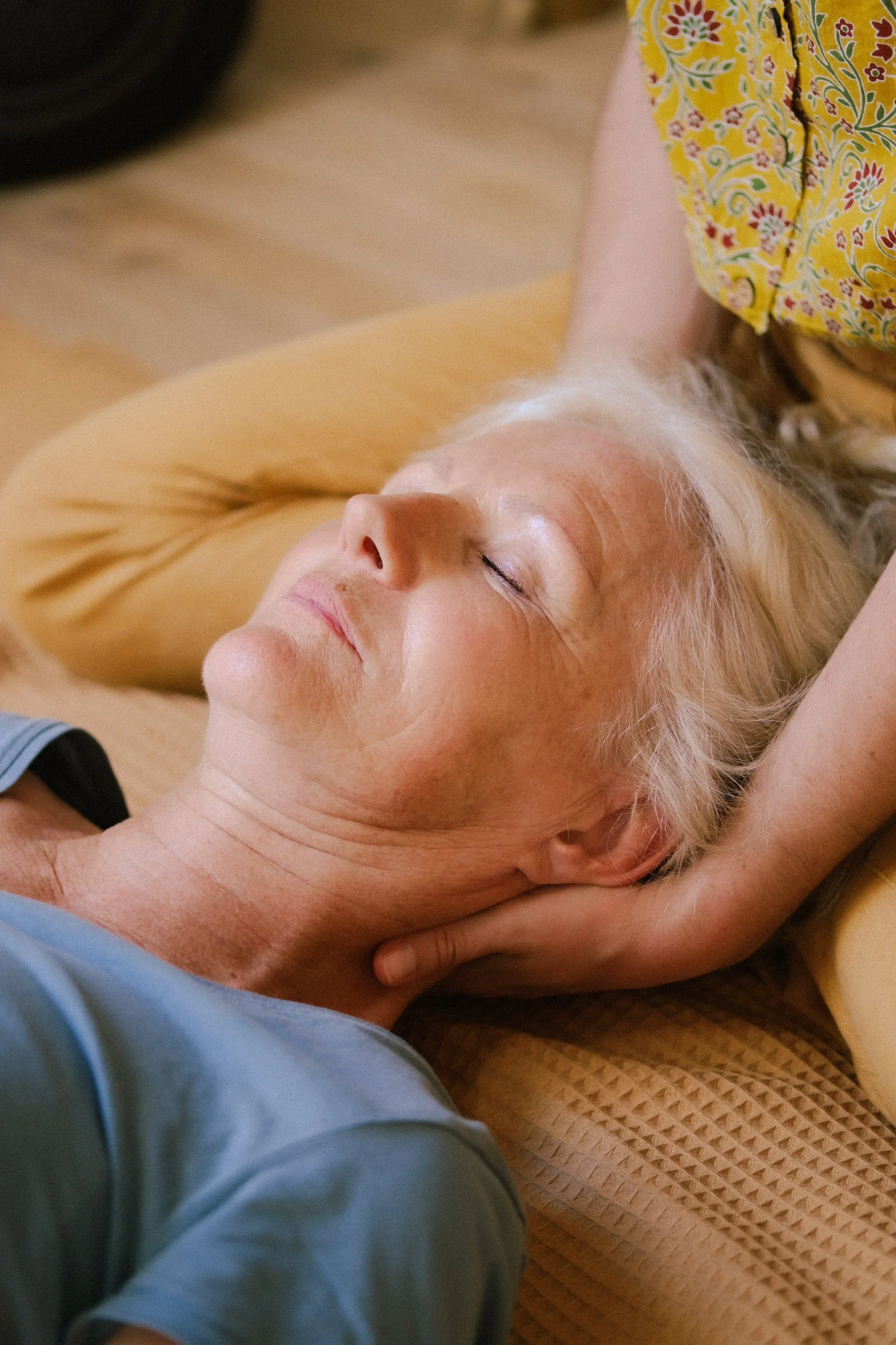 Une femme âgée avec des cheveux blonds bercés, qui a l'air détendue, repose sur un futon, avec une main derrière la tête, dans un moment de calme et de relaxation.