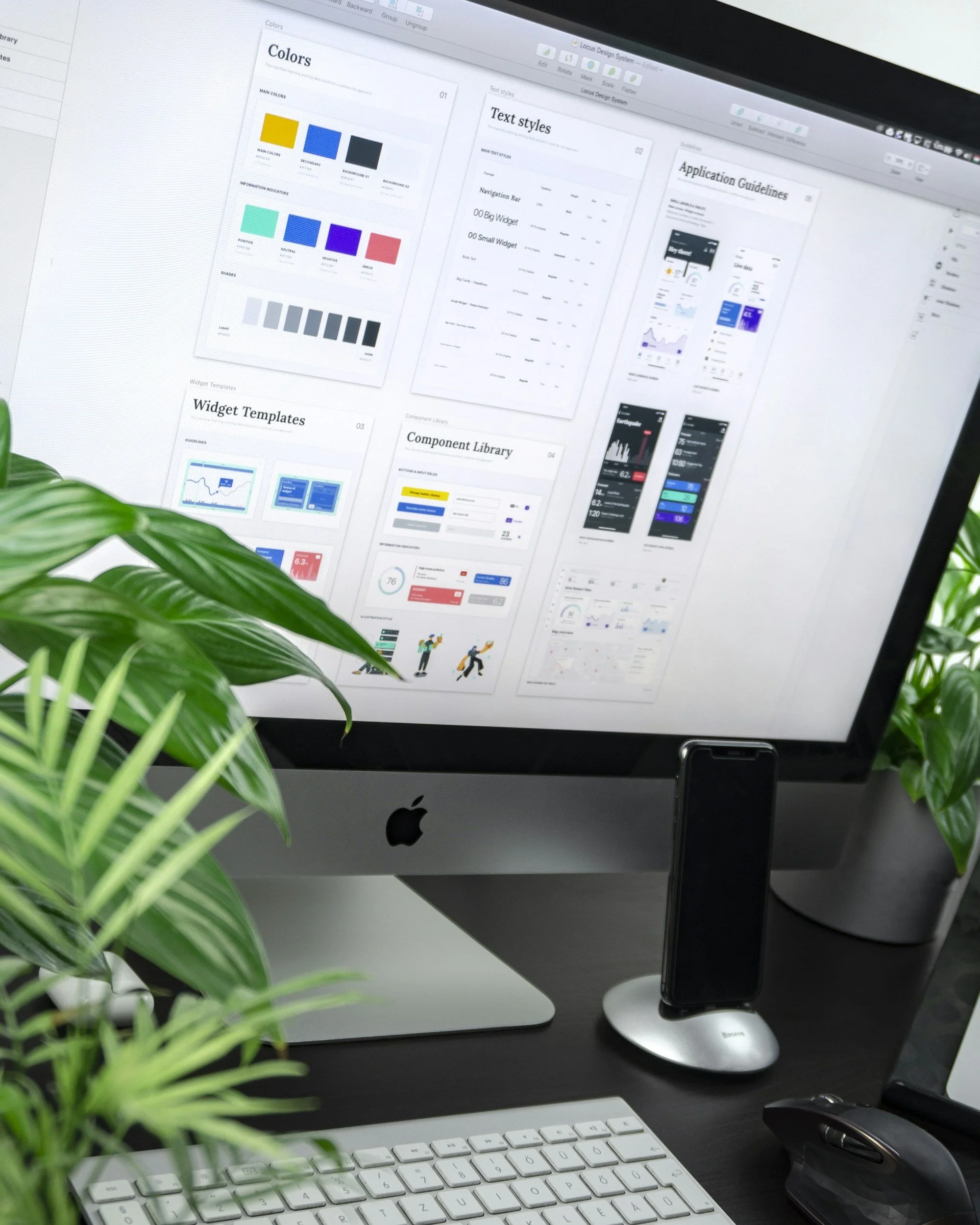 Ein Computerbildschirm zeigt verschiedene Designvorlagen für eine App, einschließlich Farbpaletten, Schriftarten und Widget-Layouts. Der Bildschirm ist auf einem Schreibtisch mit einer Tastatur, Maus, einem iPhone, einem Ständer für das iPhone und einigen grünen Pflanzen im Vordergrund zu sehen.