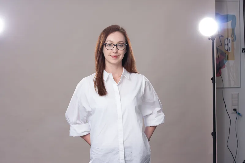 Frau mit Brille und weißen Hemd posiert in einem Fotostudio vor neutralem Hintergrund.