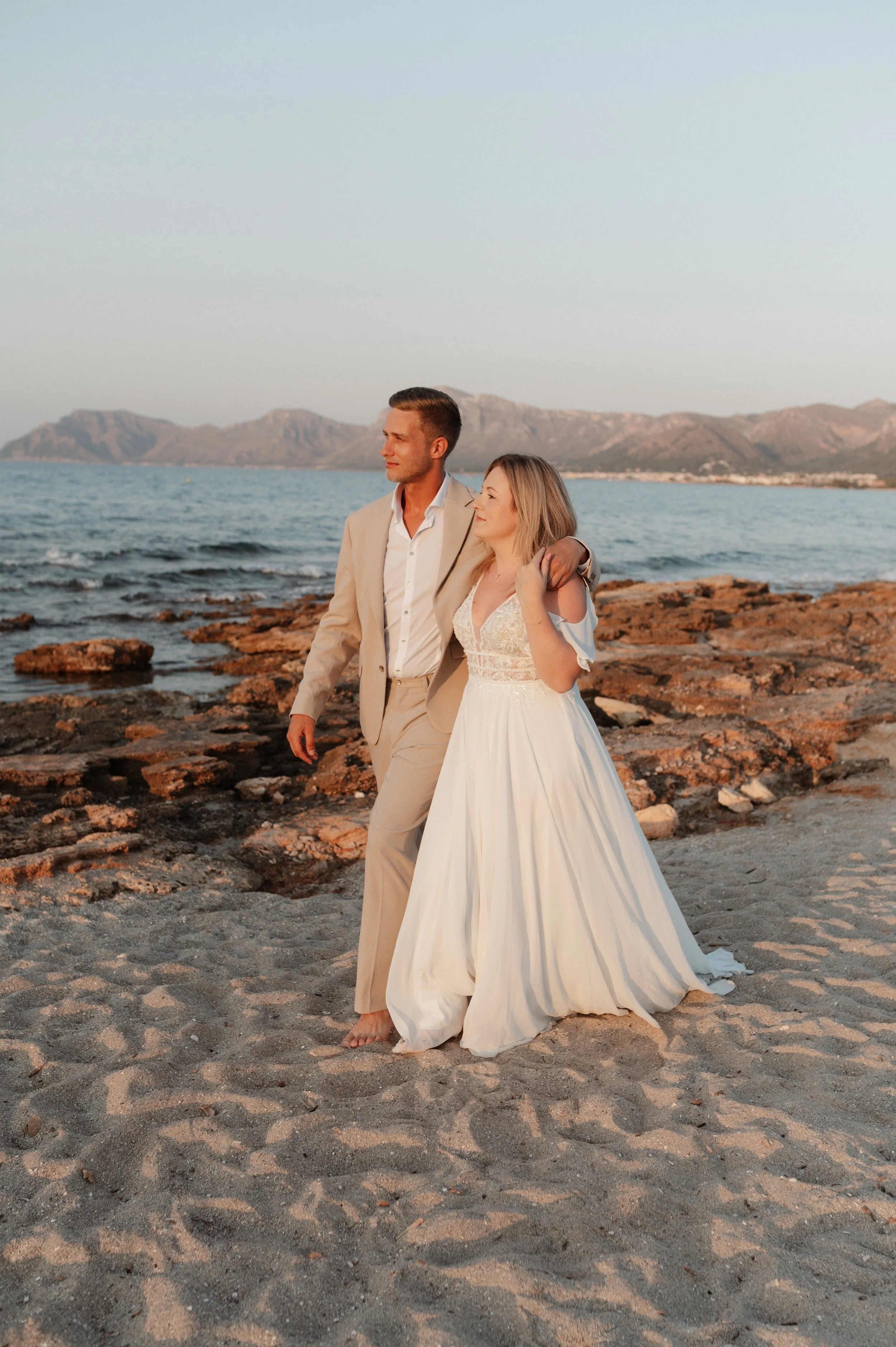Ein Paar in Hochzeitskleidung am Strand bei Sonnenuntergang, im Hintergrund Berge und das Meer.