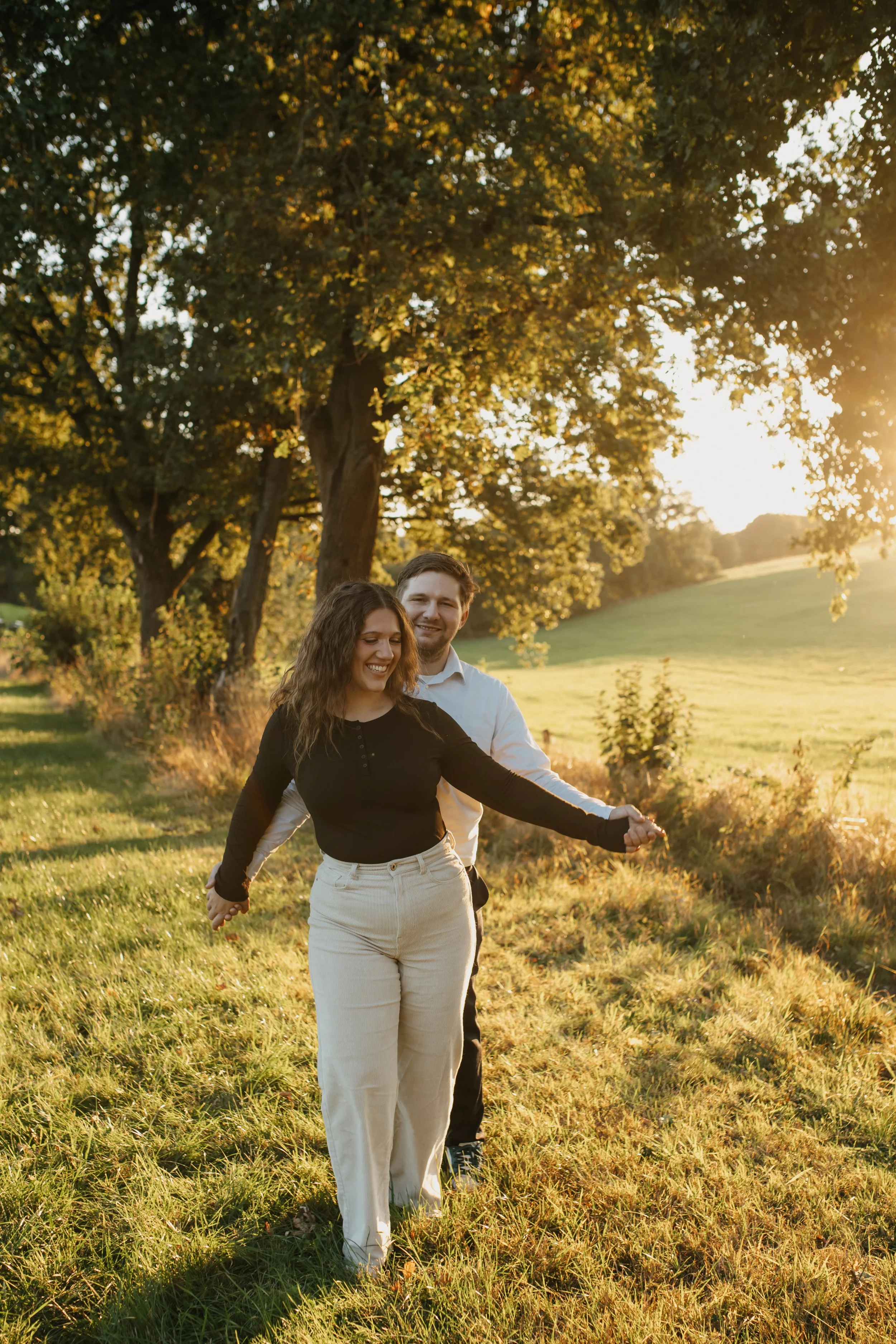 Ein Paar, ein Mann und eine Frau, tanzen Hand in Hand in einem sonnigen Park bei Sonnenuntergang, umgeben von Bäumen und grünem Gras.
