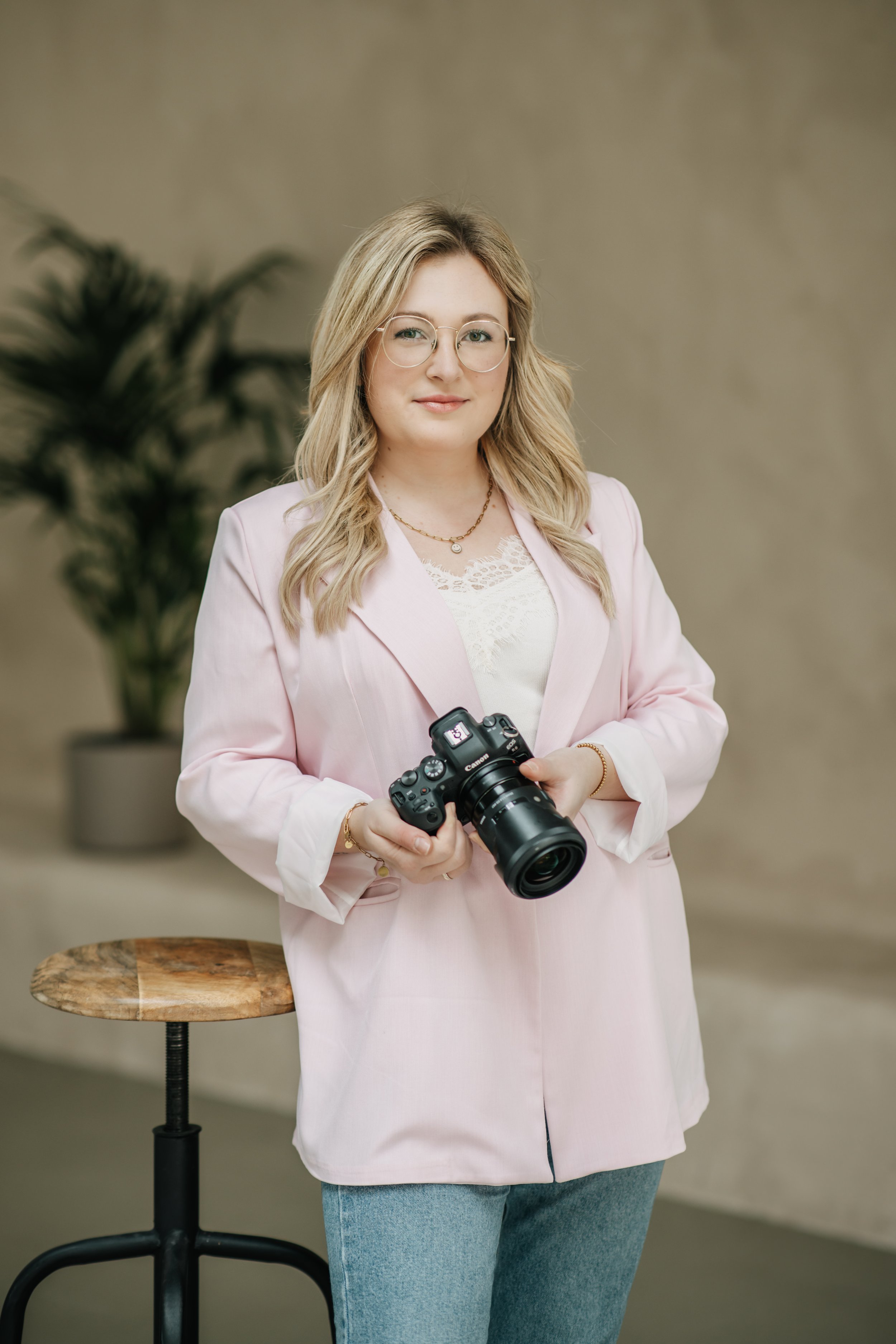 Eine Frau mit blondem Haar, Brille, rosa Jacke und Jeans hält eine Kamera in den Händen, im Hintergrund eine Pflanze