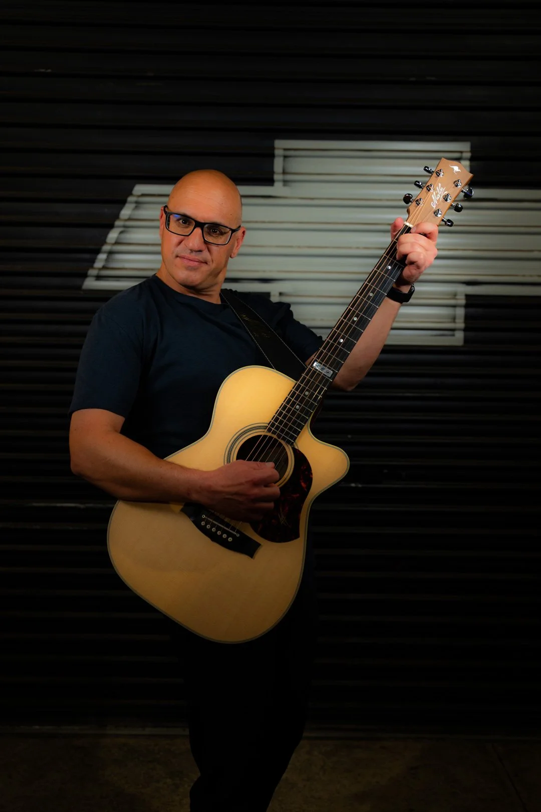A man with glasses holding an acoustic guitar in front of a black and white background with musical staff design.