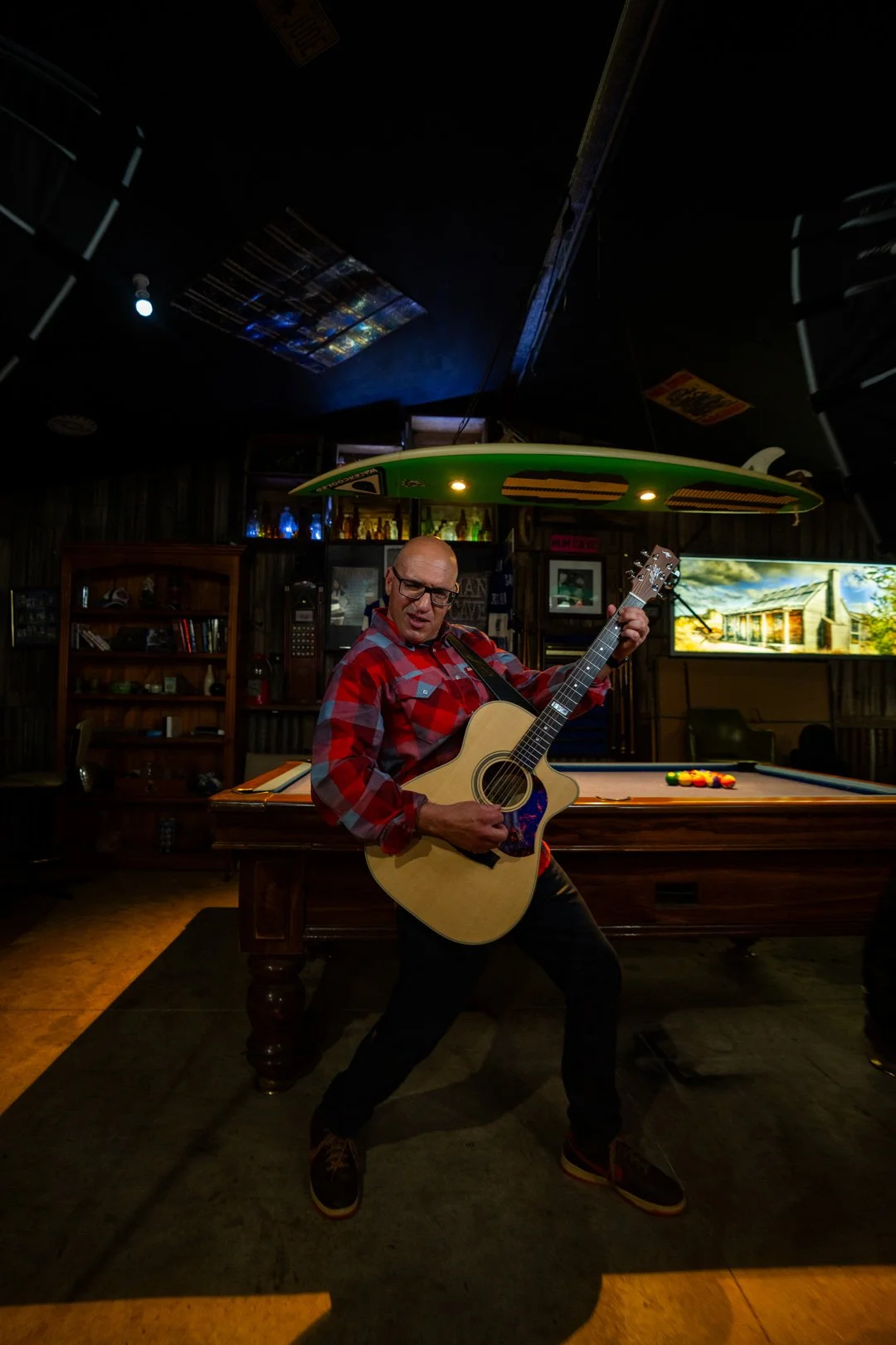 A man wearing glasses and a red plaid shirt playing an acoustic guitar in a dimly lit bar or lounge with a pool table and neon signs in the background.