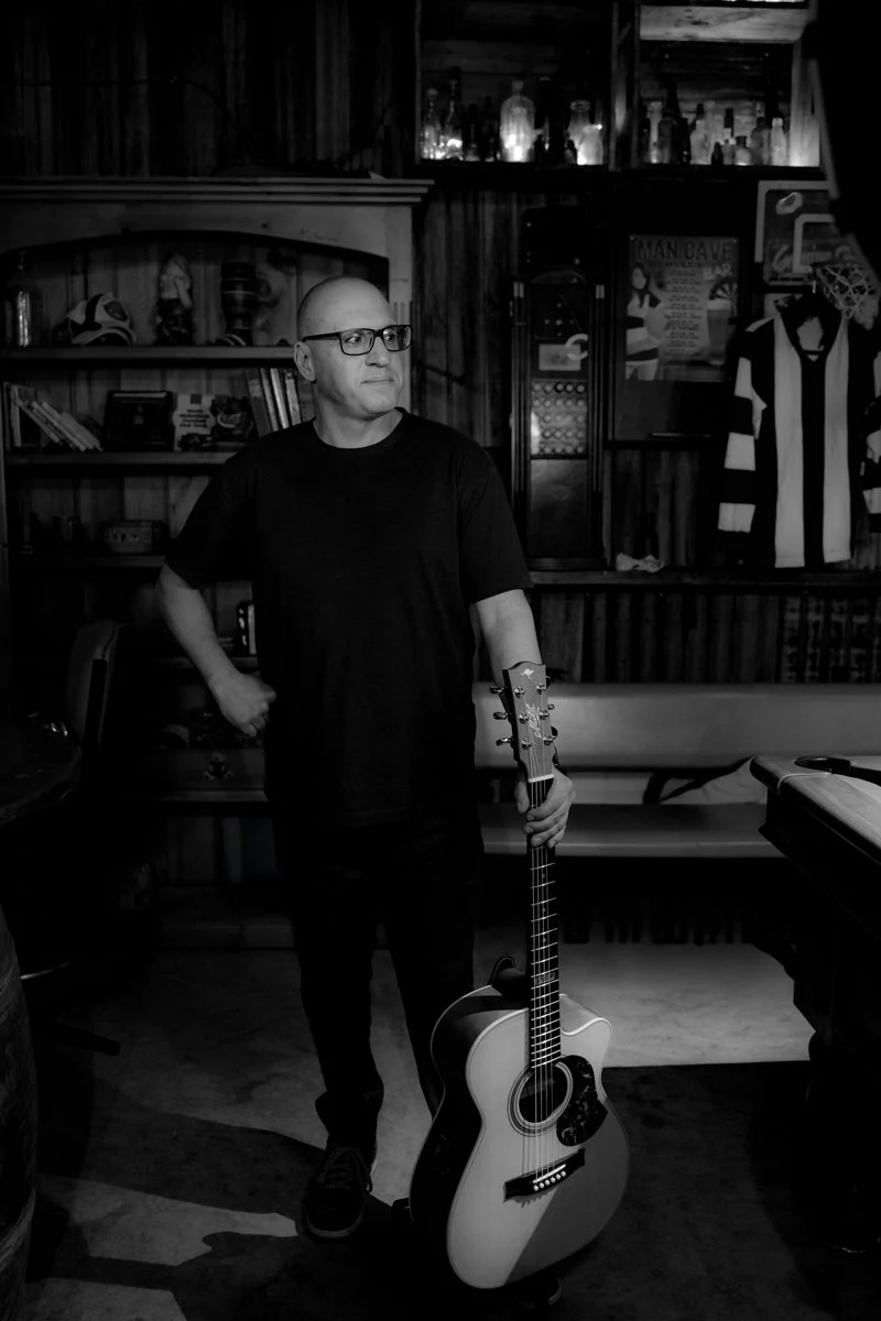 A man with glasses standing in a bar or cafe, holding an acoustic guitar, surrounded by shelves with books and bottles, and sports jerseys on the wall, in black and white.