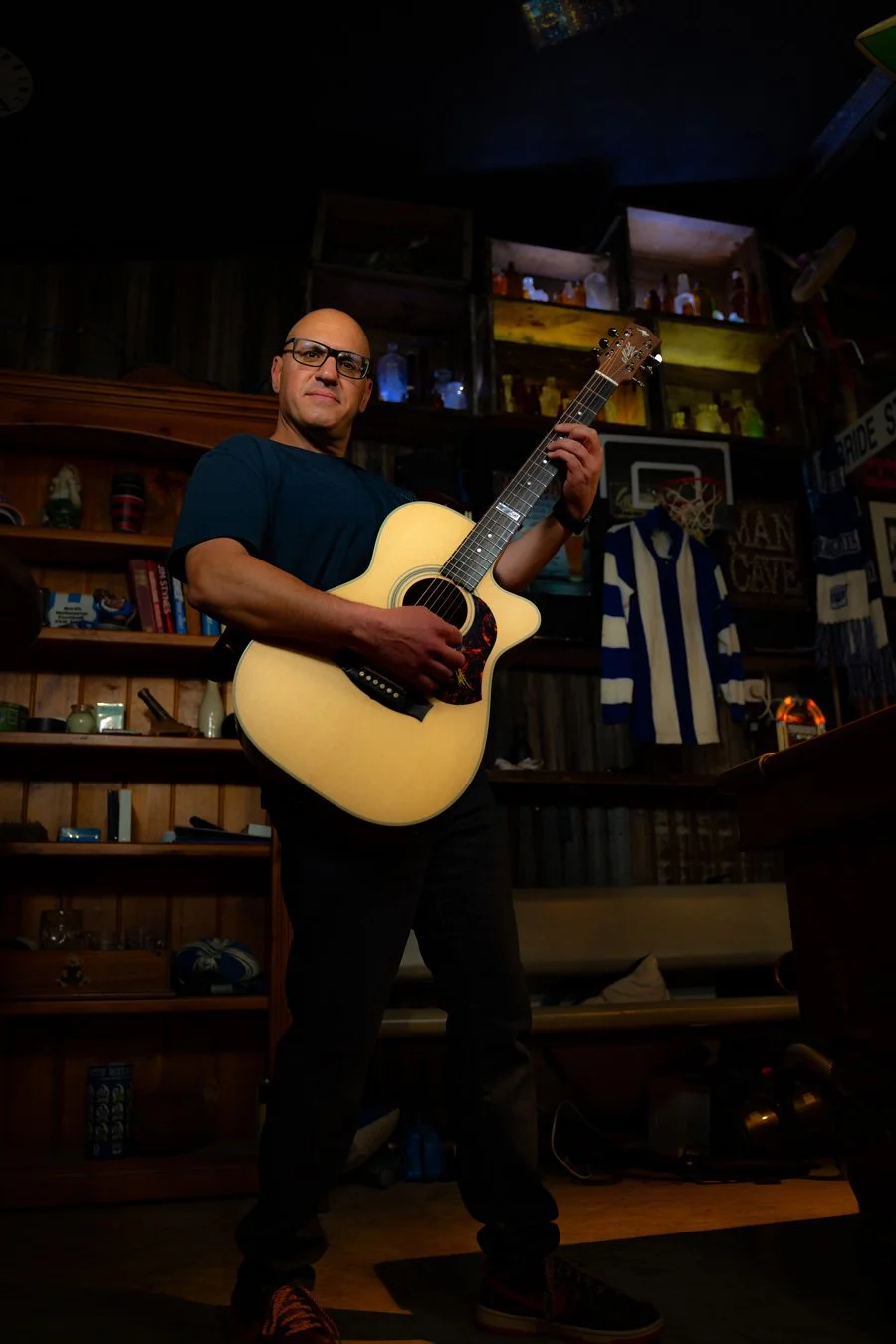 A man wearing glasses and a dark t-shirt playing an acoustic guitar in a dimly lit room with wooden shelves and sports memorabilia.