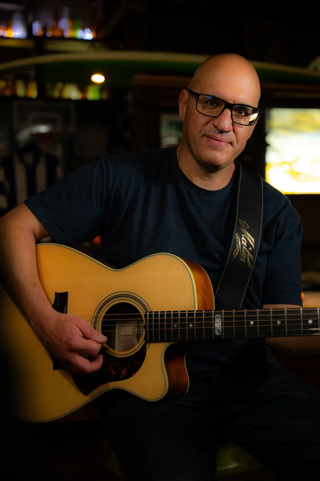 A man with glasses playing an acoustic guitar in a dimly lit room.