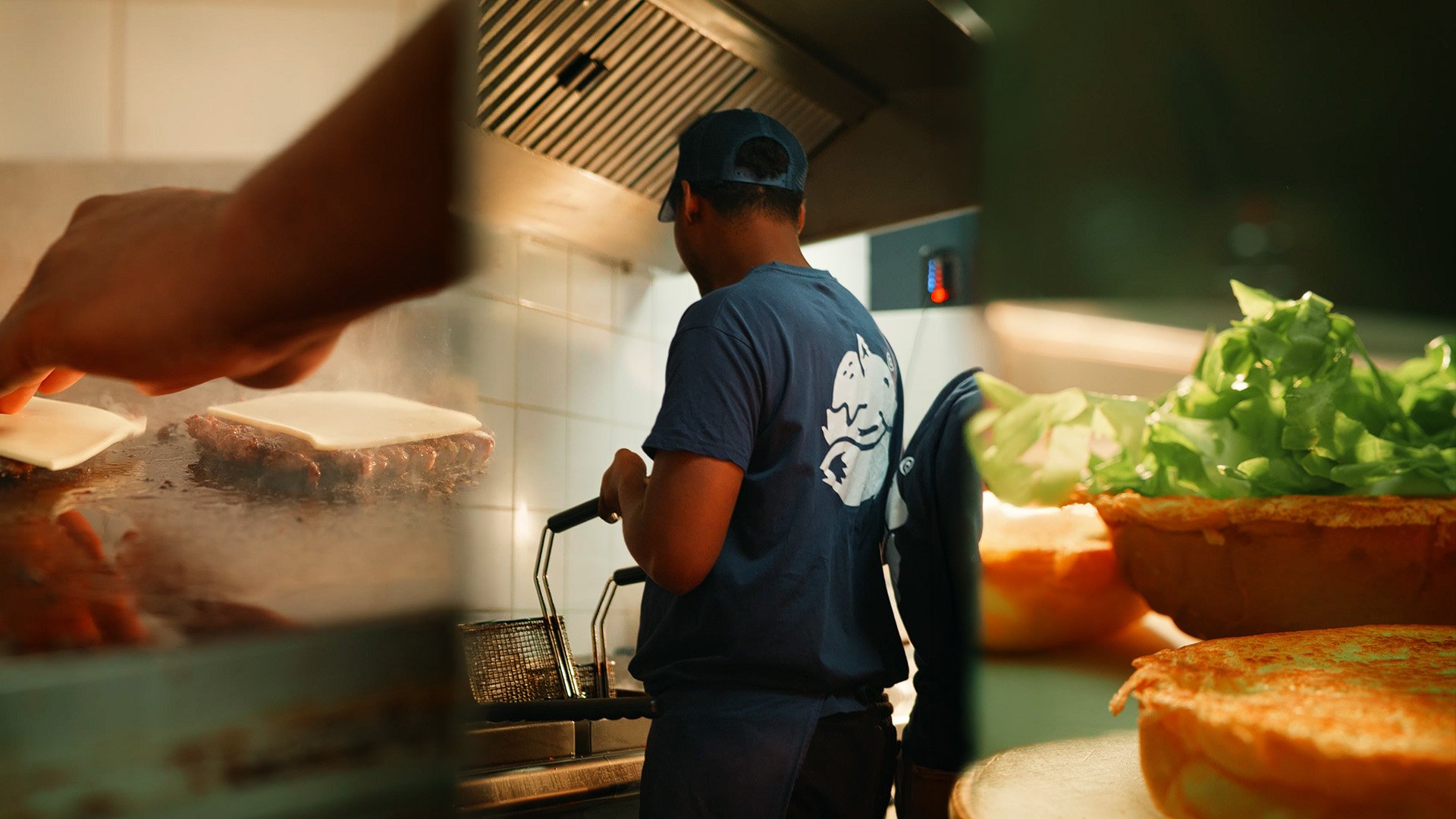 Un homme portant un t-shirt bleu et une casquette noire cuisine dans une cuisine de restaurant, avec une plaque de cuisson professionnelle et des légumes verts sur du pain à burger.