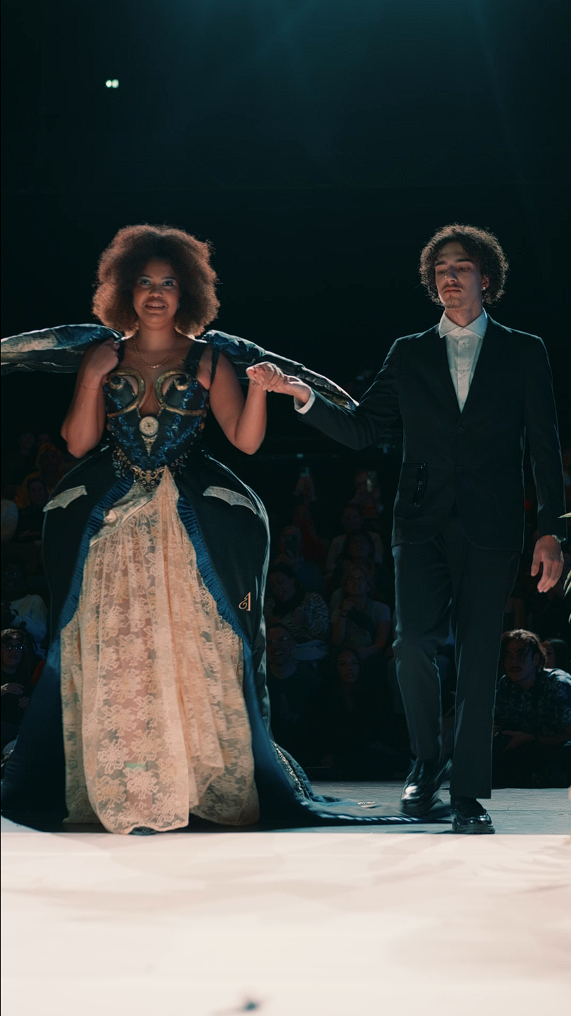 Un couple défile lors d'une représentation de mode, la femme porte une robe spectaculaire avec un corset décoratif, et l'homme en élégant costume noir lui tient la main pendant le défilé.