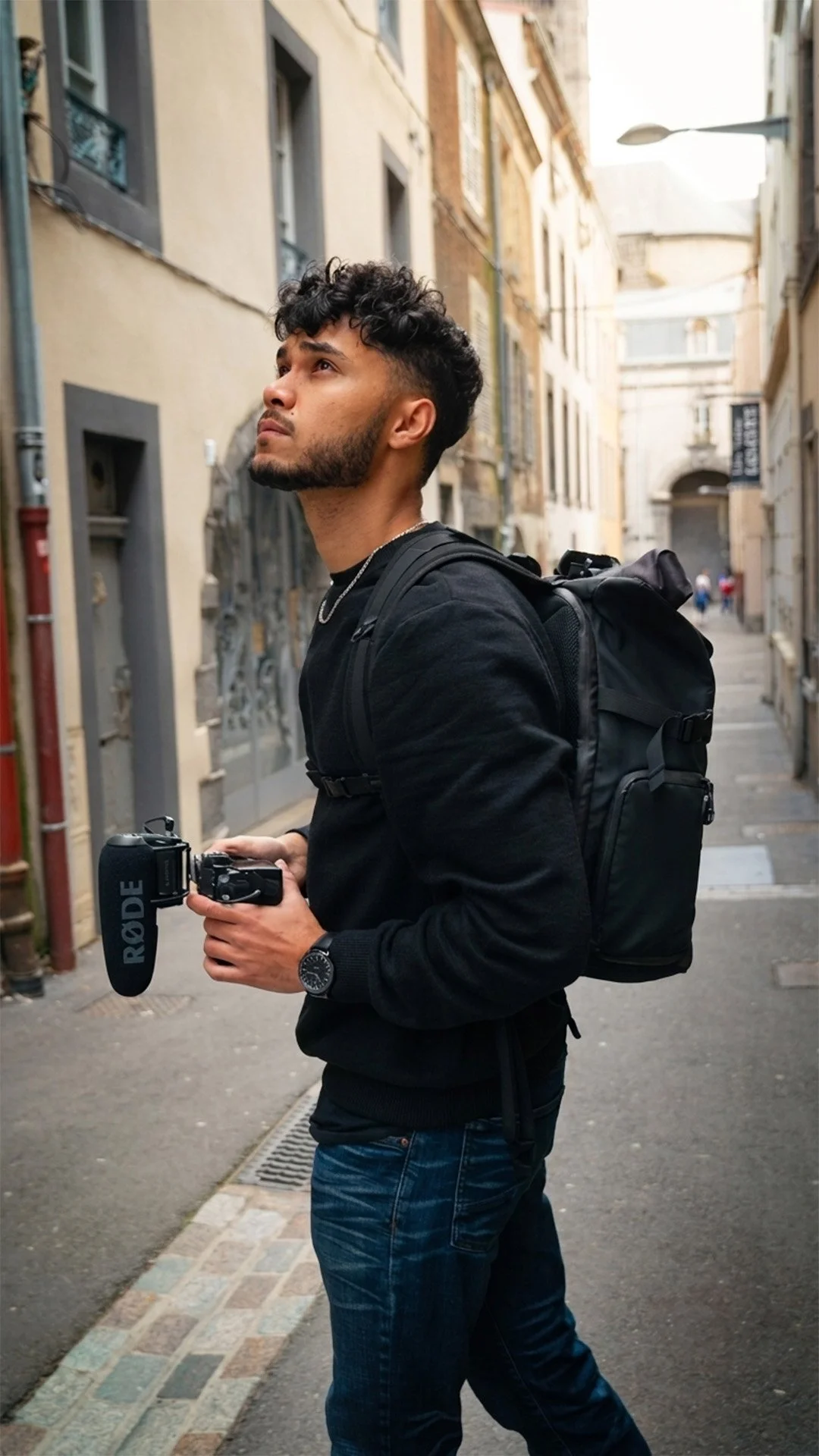 Un homme avec une sac à dos noir tient une caméra avec un microphone Rode dans une rue pavée, en arrière-plan des bâtiments anciens en pierre et une lanterne suspendue.