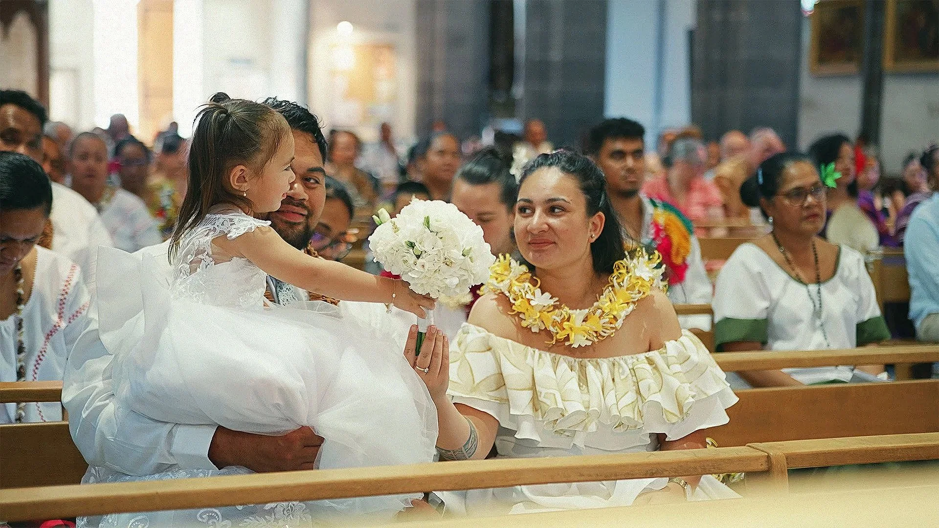 Une cérémonie religieuse avec un homme portant une petite fille en robe blanche qui lui donne un bouquet de fleurs, une femme portant un collier de fleurs regarde la petite fille, présence de plusieurs personnes assises dans une église