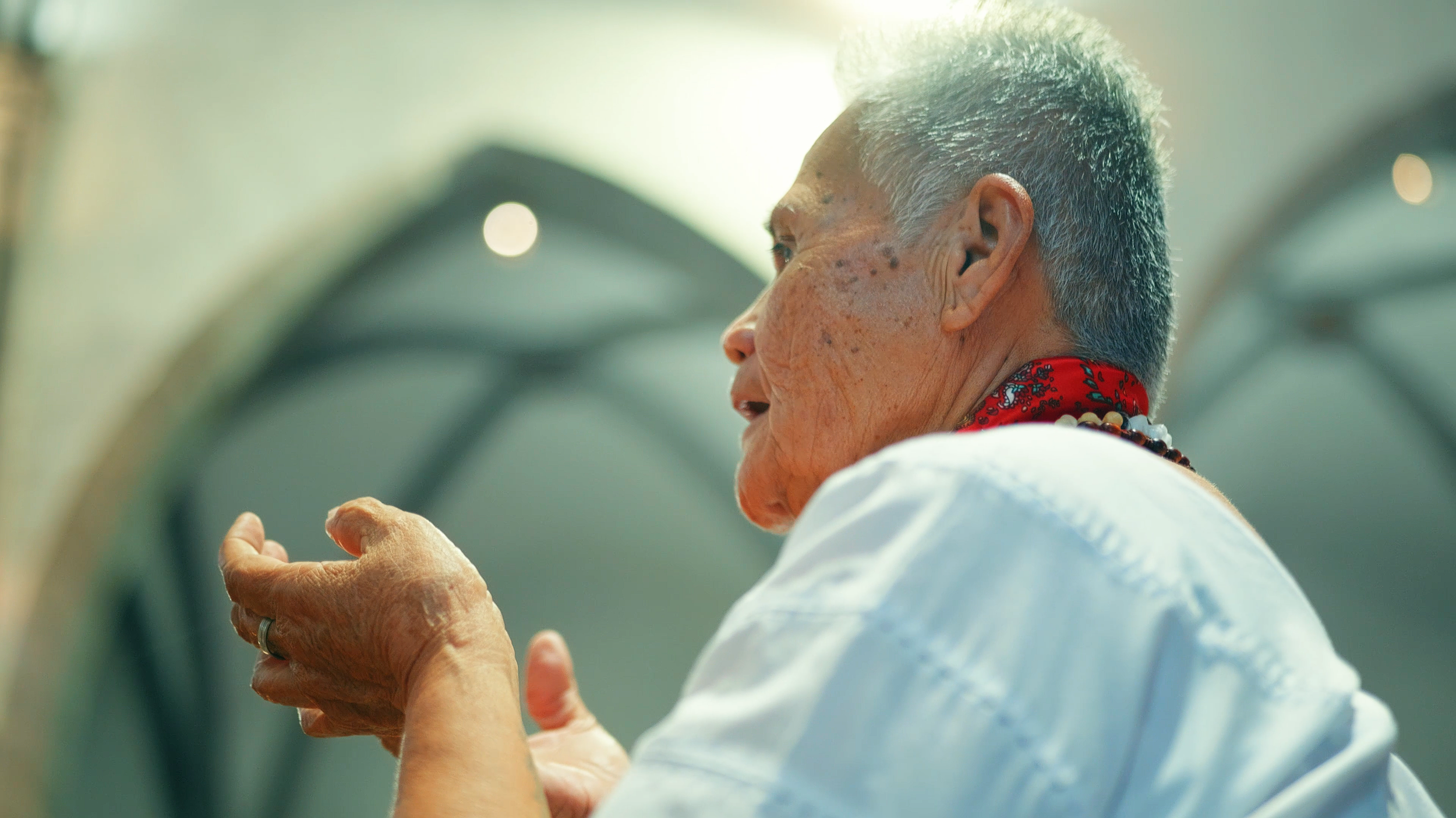Une femme âgée avec des cheveux gris, portant une robe blanche, un collier et une écharpe rouge, prie avec les mains jointes dans une église chaleureusement éclairée.