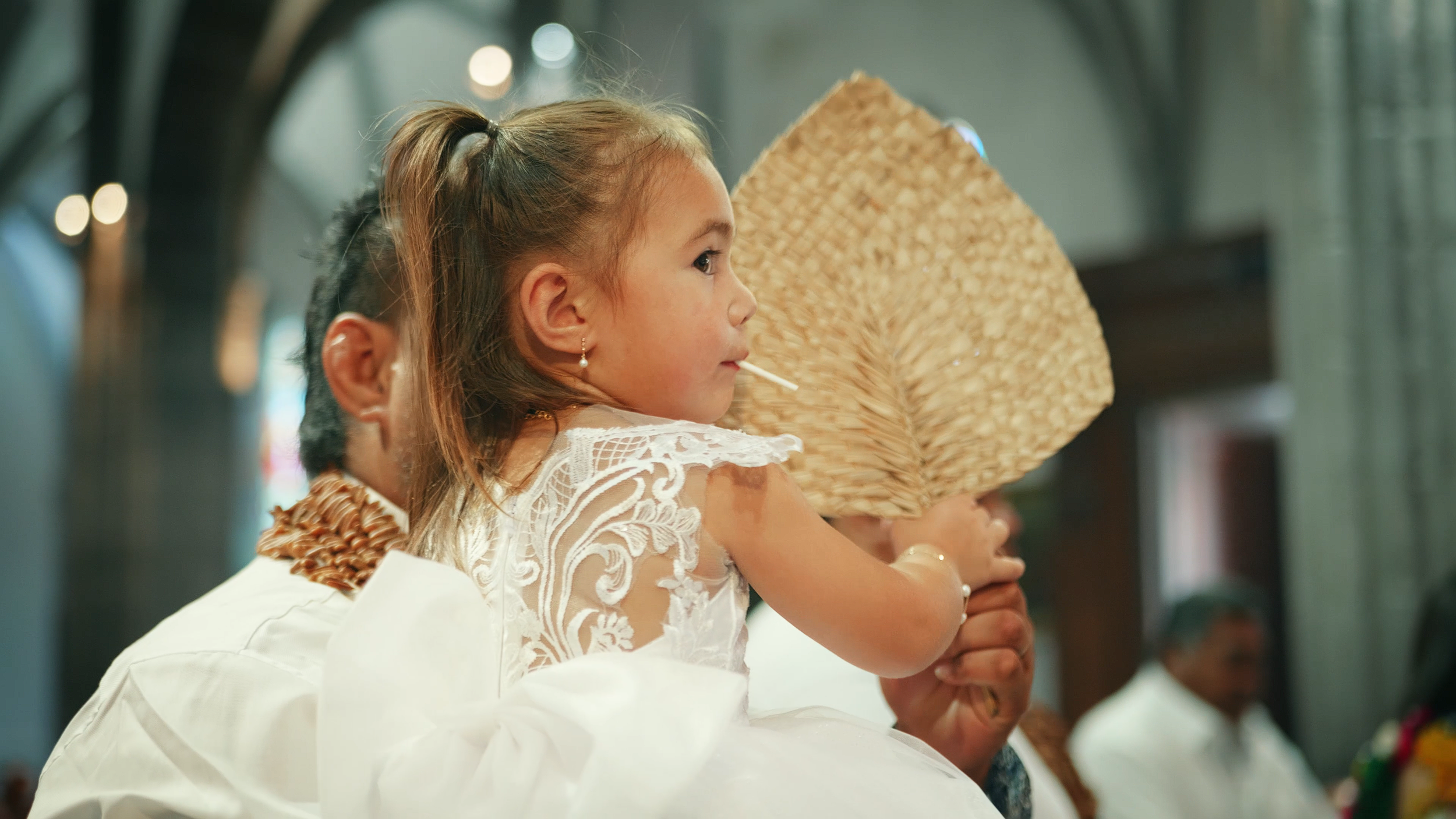 Une jeune fille avec une robe blanche en dentelle et une boucle d'oreille, qui pose avec un éventail  en feuille de cocotier, dans un contexte probablement cérémonial, en arrière-plan un homme portant un vêtement blanc et un collier, dans une église.