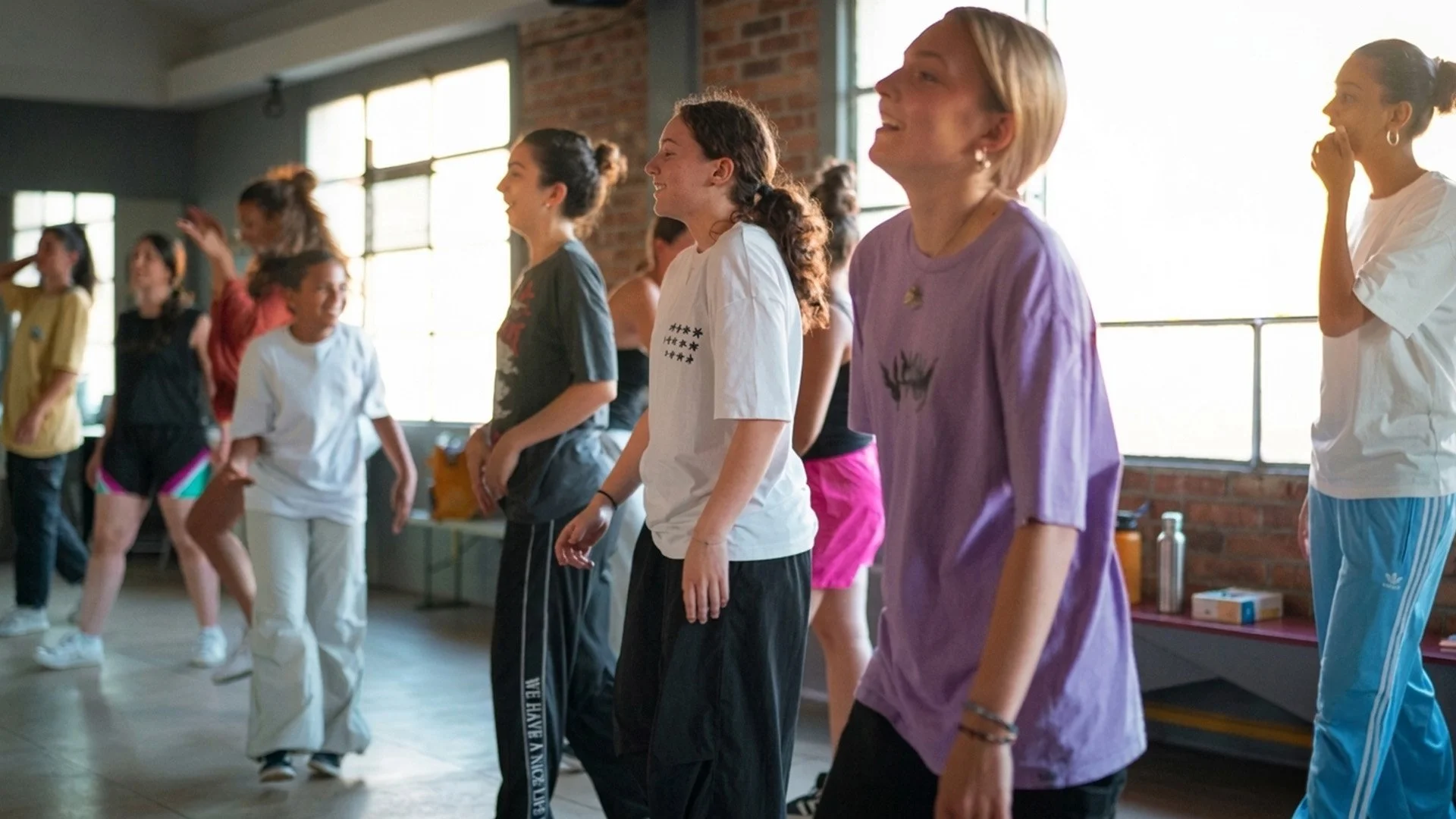 Groupe de personnes, principalement des femmes et filles adolescentes, en train de danser ou de faire une activité de groupe dans un studio avec des murs en briques et de grandes fenêtres.