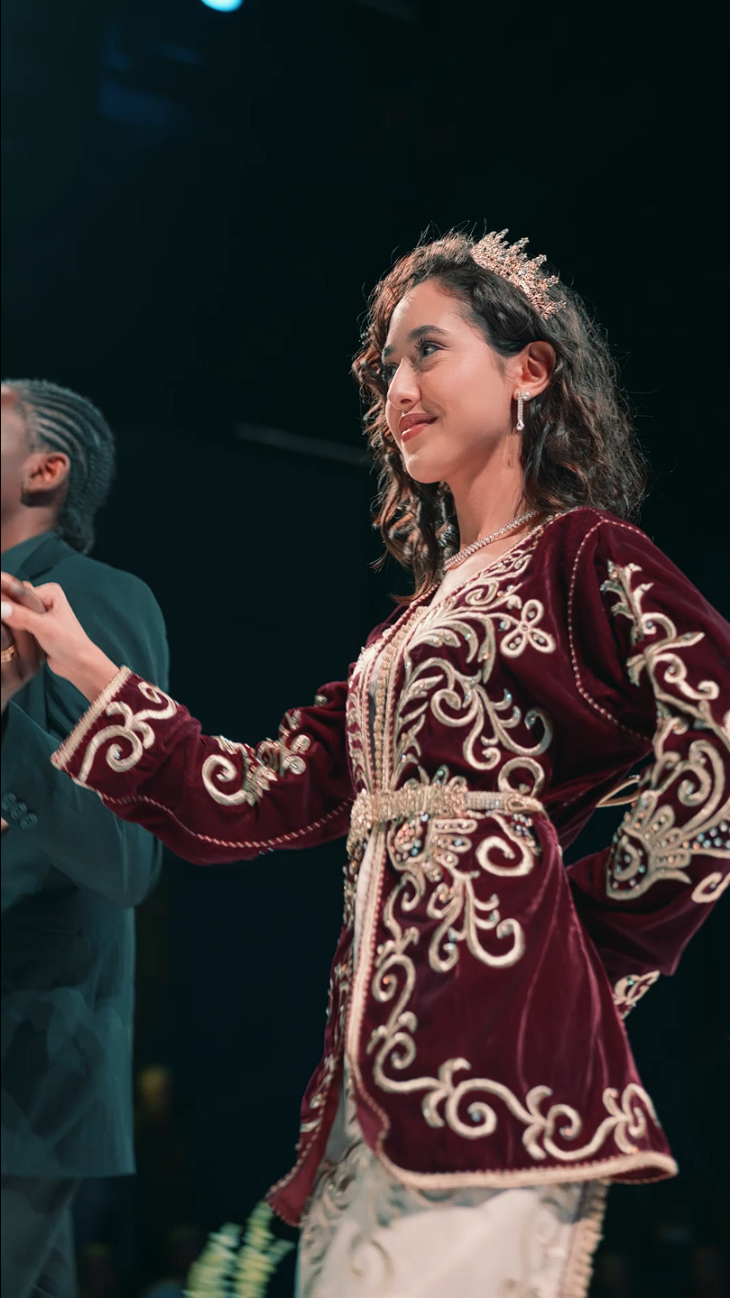 Une jeune femme portant une robe de cérémonie rouge ornée de broderies dorées, un diadème, et des bijoux, lors d'un grand défilé de mode.