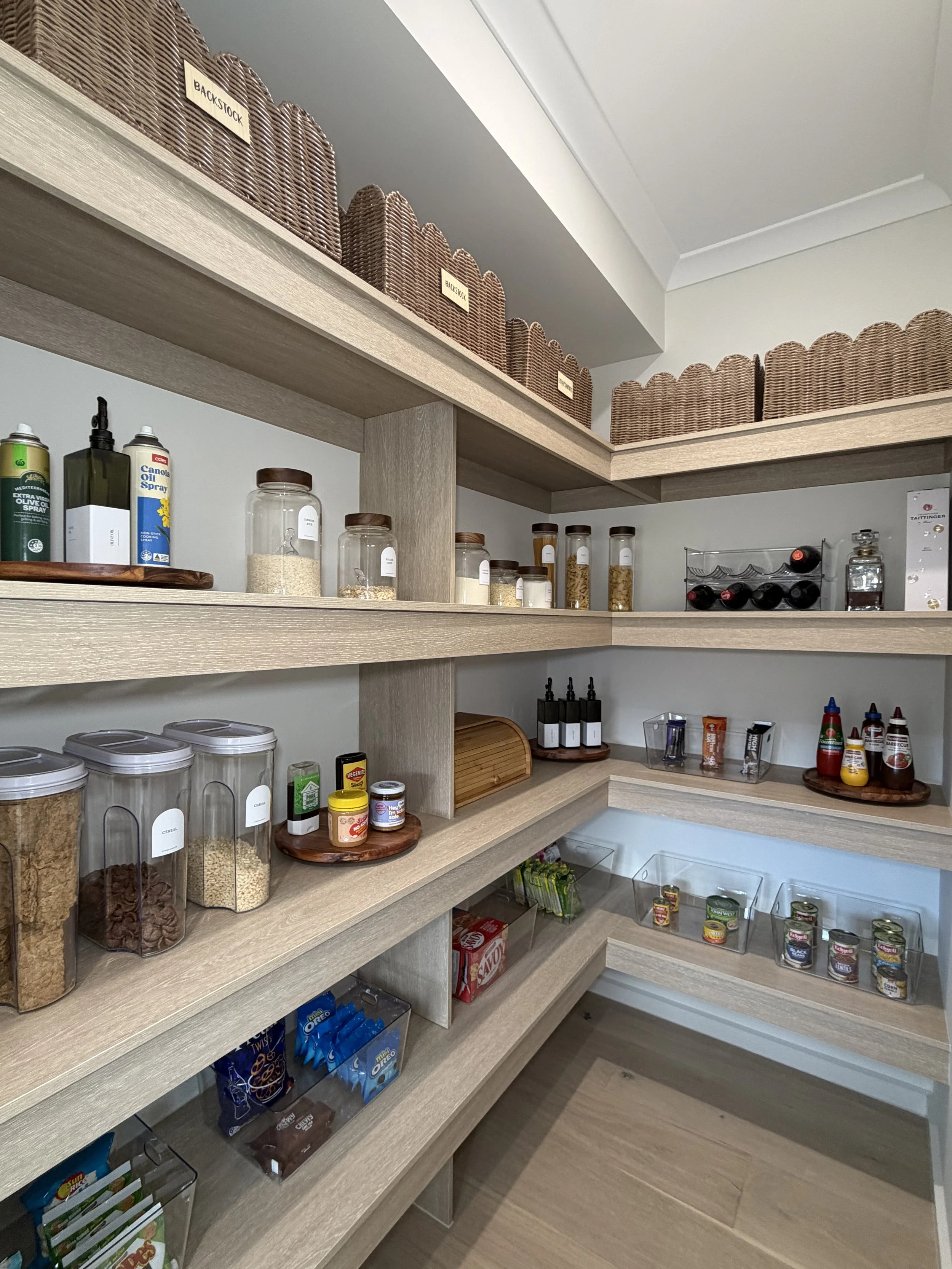 Walk-in pantry with wooden shelving holding woven baskets, jars and bottles.