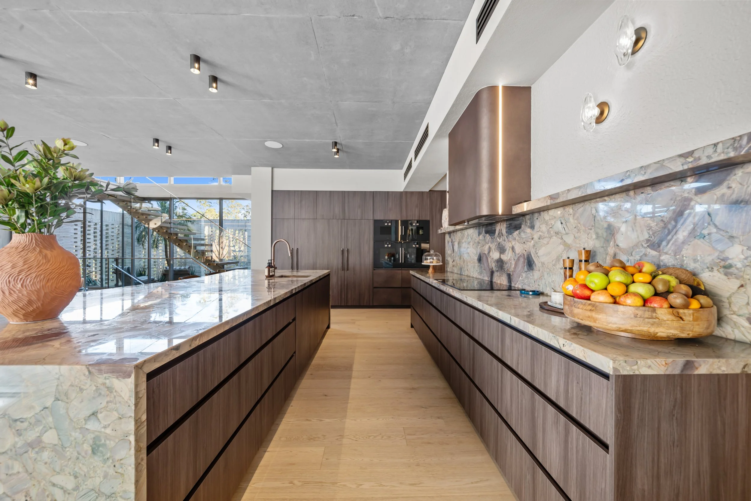 Long galley kitchen at Stella Place with dark timber cabinetry, stone benchtops and a window view.