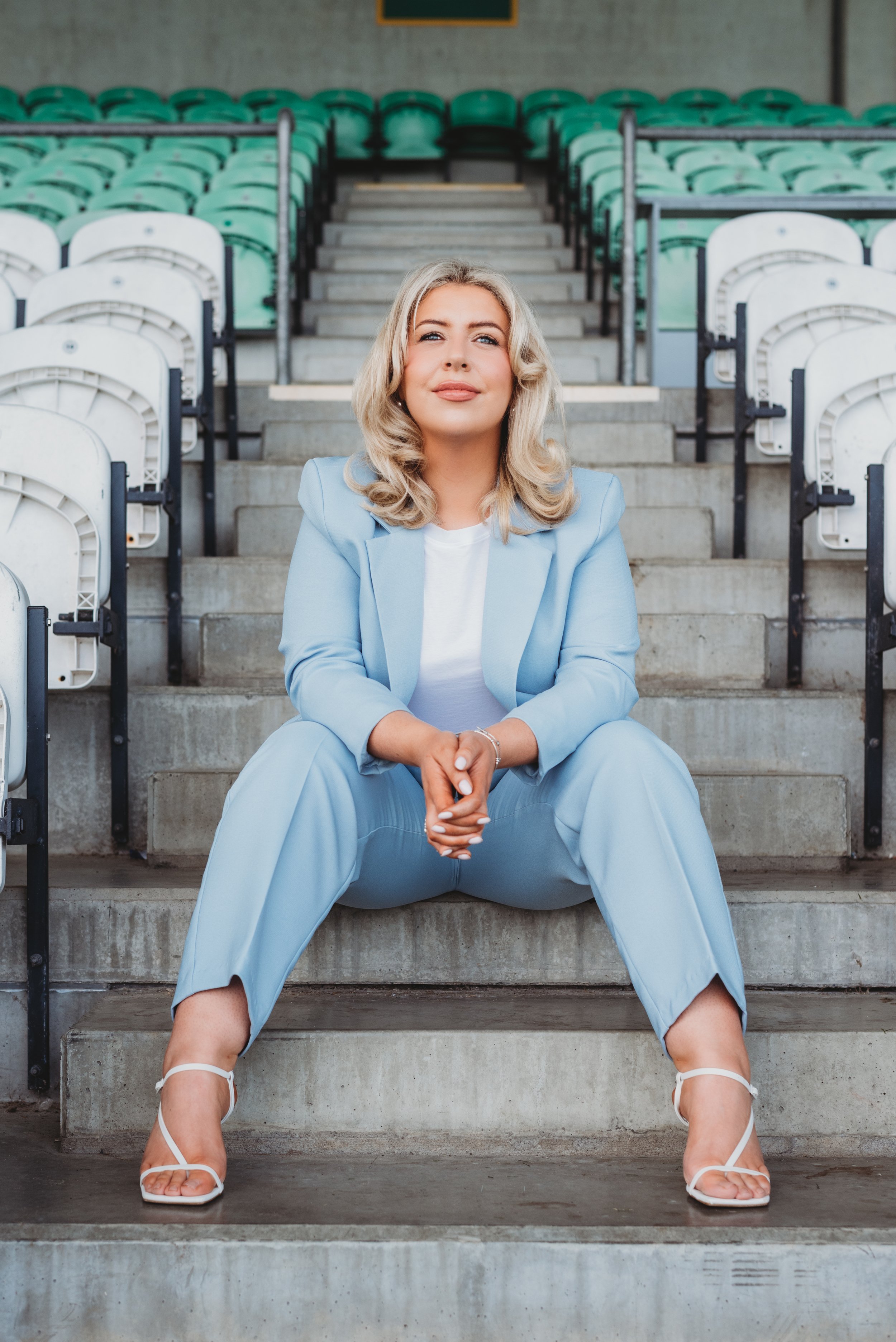 A woman with blonde curly hair sitting on concrete stadium stairs, wearing a light blue suit and white sandals.