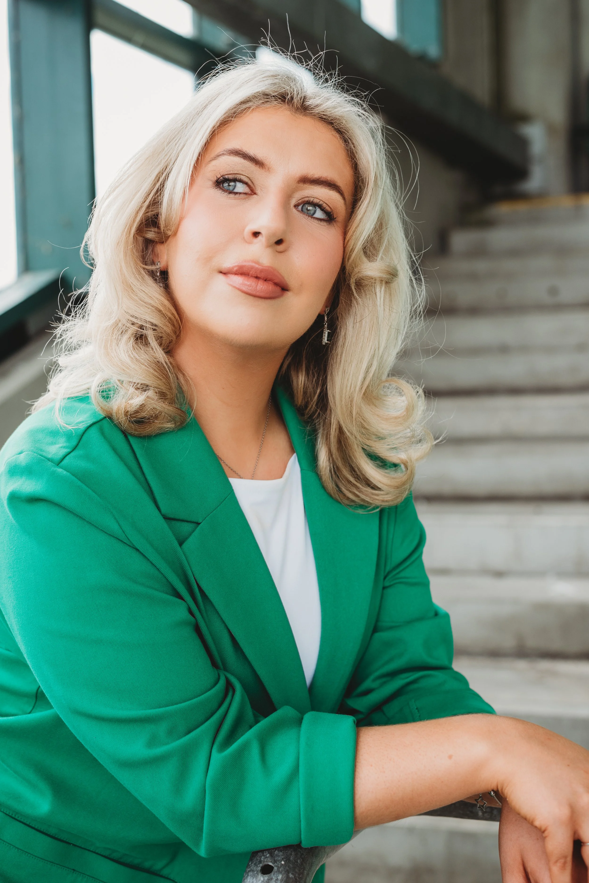 Close-up of a woman with blonde, wavy hair, blue eyes, wearing a green blazer and white shirt, sitting on steps with her arms crossed, looking to her left, with a window and staircase in the background.