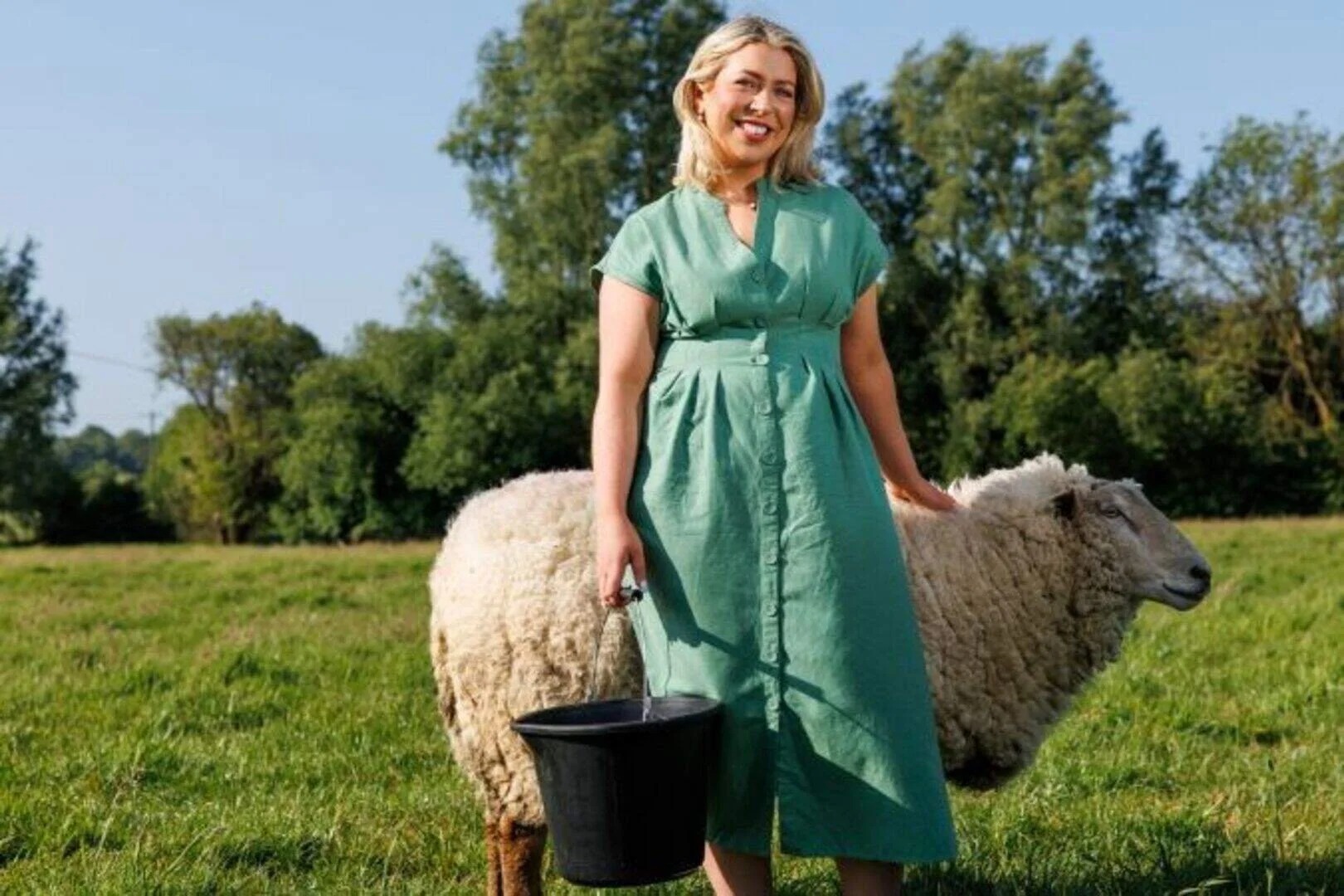 A woman in a green dress standing outdoors on a farm, holding a black bucket, with a sheep grazing next to her and trees in the background.