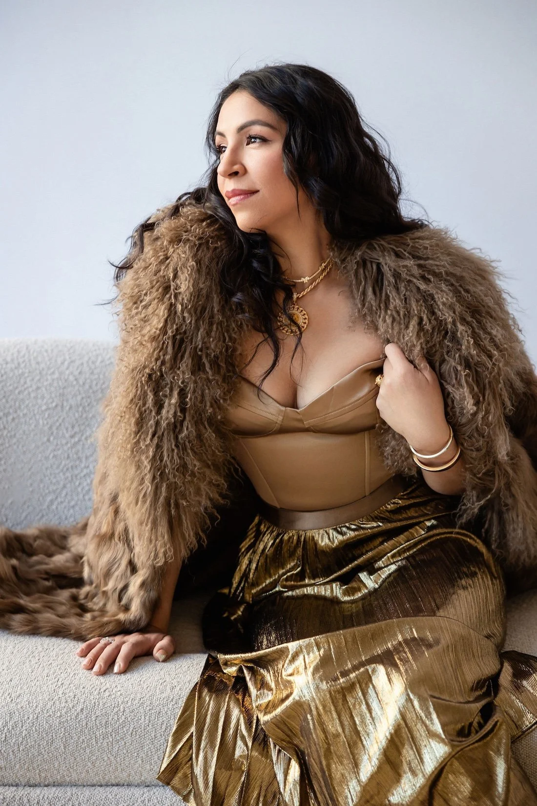 A woman with dark, curly hair and light skin, dressed in a shiny gold and black skirt, a beige bustier top, and a brown fur coat, sitting on a light-colored sofa.