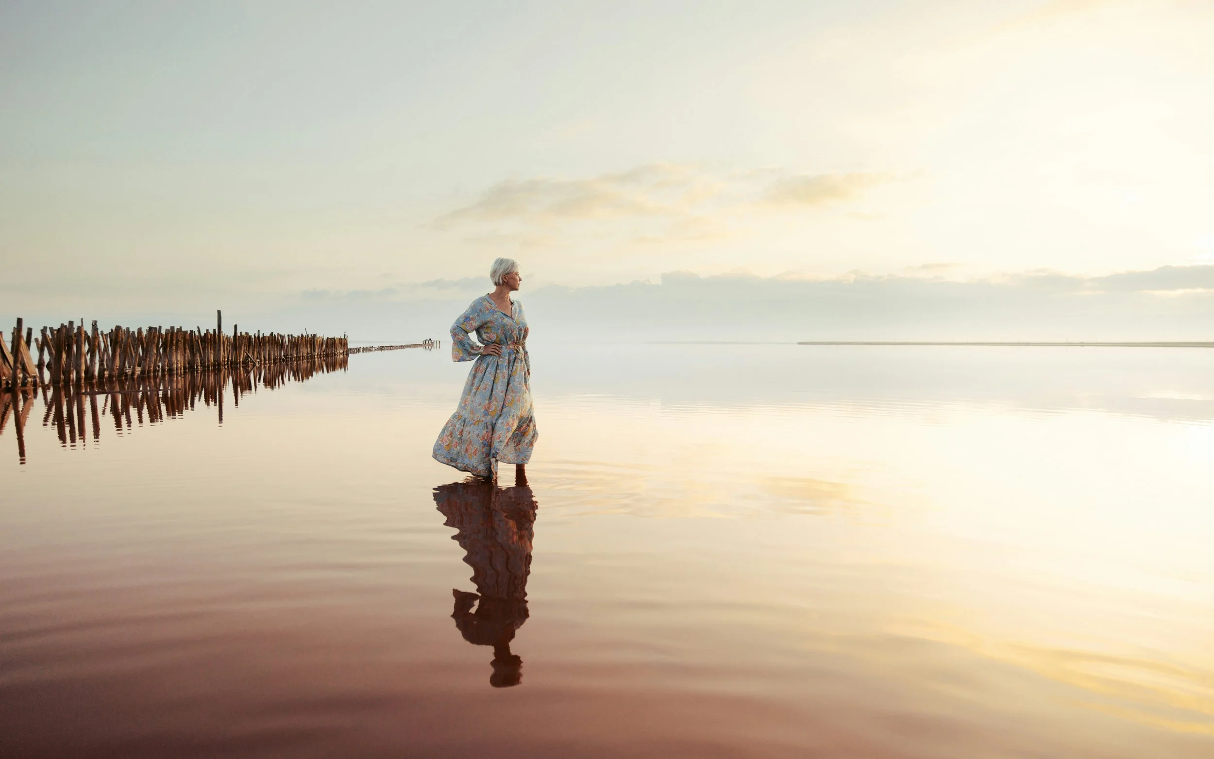 A woman with gray hair standing in shallow water at sunset or sunrise, wearing a colorful dress with a floral pattern, facing left with hands on hips, with a wooden pier visible in the background.